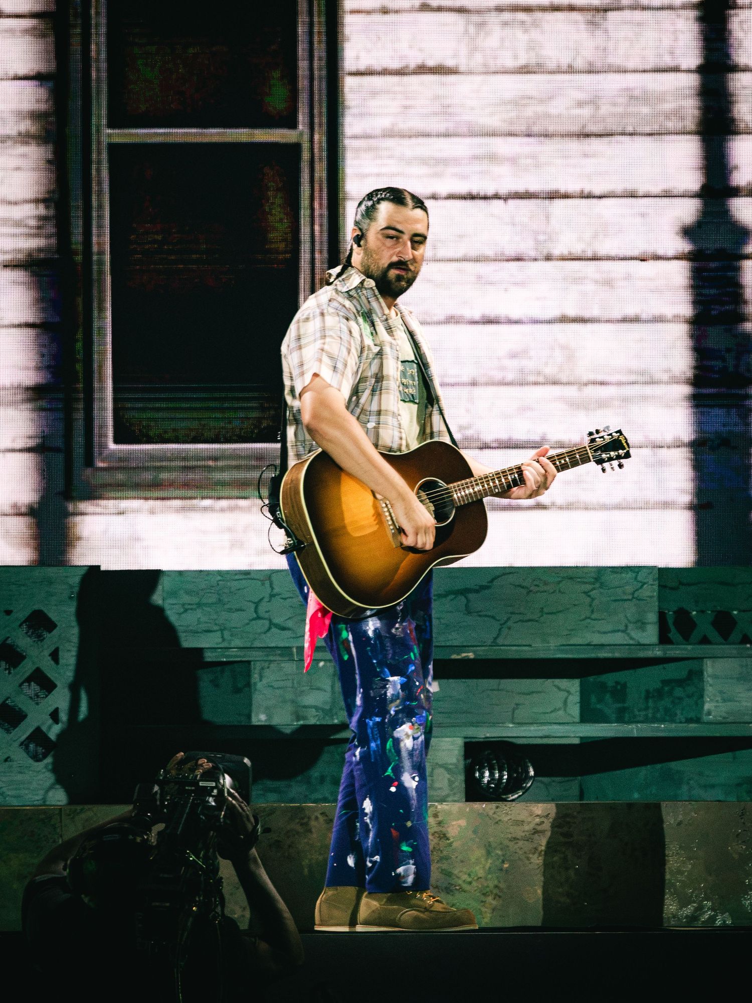 Bearded male musician holding acoustic guitar on stage wearing plaid shirt and colorful floral pants with house backdrop.