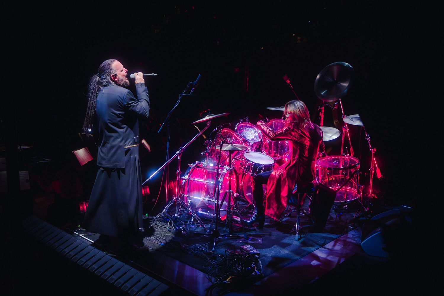 A vocalist in a long coat performs on stage beside a drummer with a glowing red drum kit in a dark concert venue.
