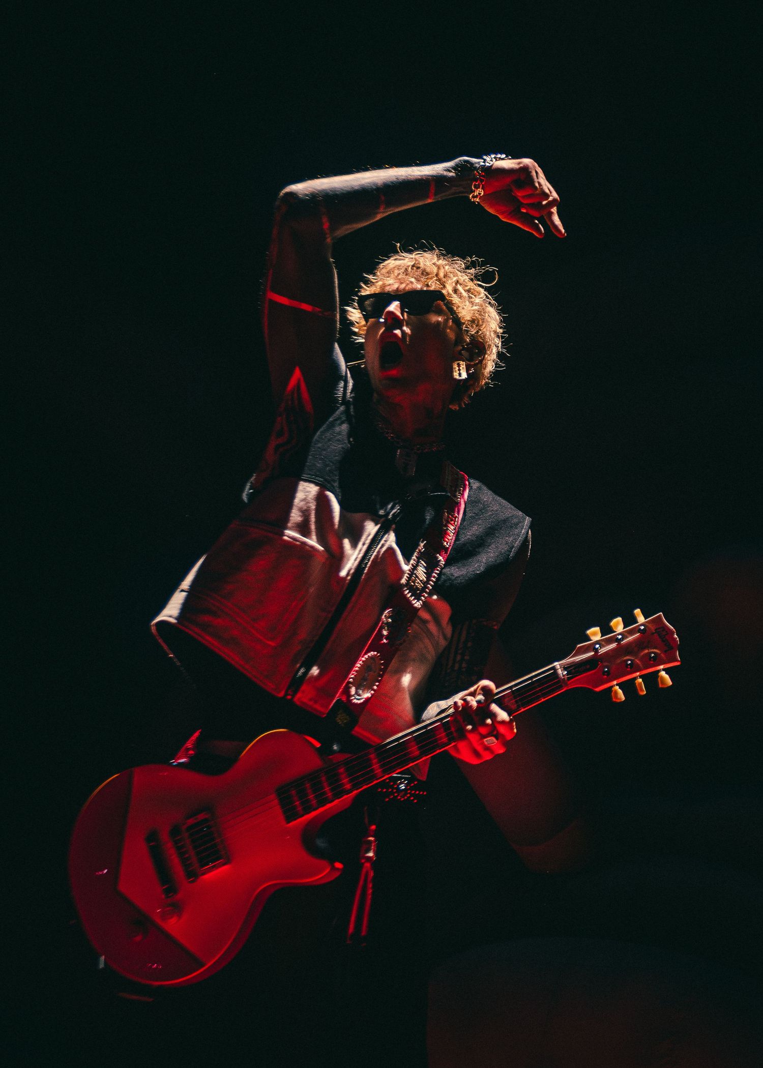 A rock guitarist leaps energetically on stage, playing a red electric guitar under dramatic red lighting.