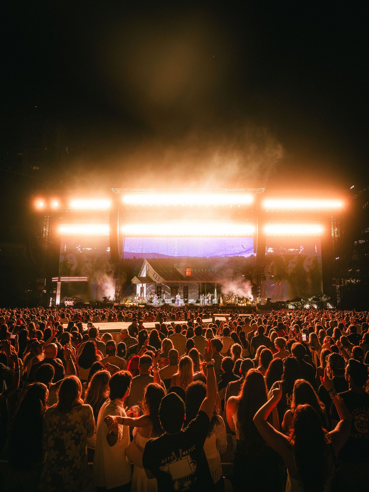 A massive outdoor concert crowd illuminated by bright orange stage lights at night.