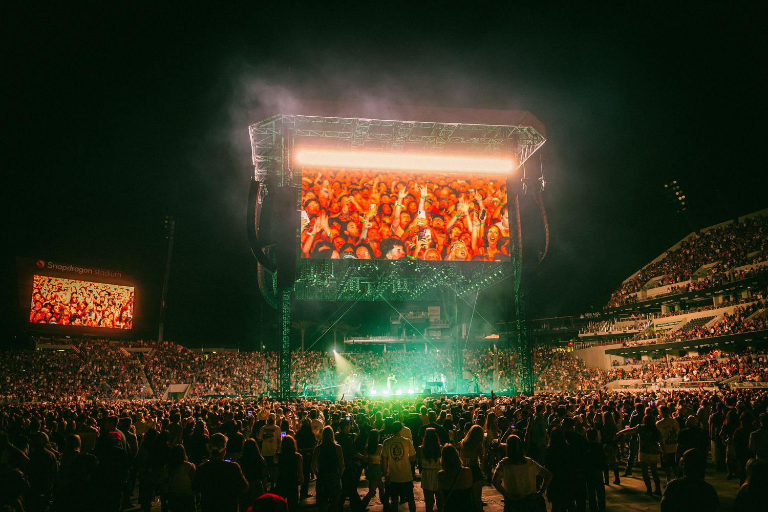 Massive outdoor concert at night with glowing green stage lights, giant LED screen, and thousands of fans packed in the crowd.