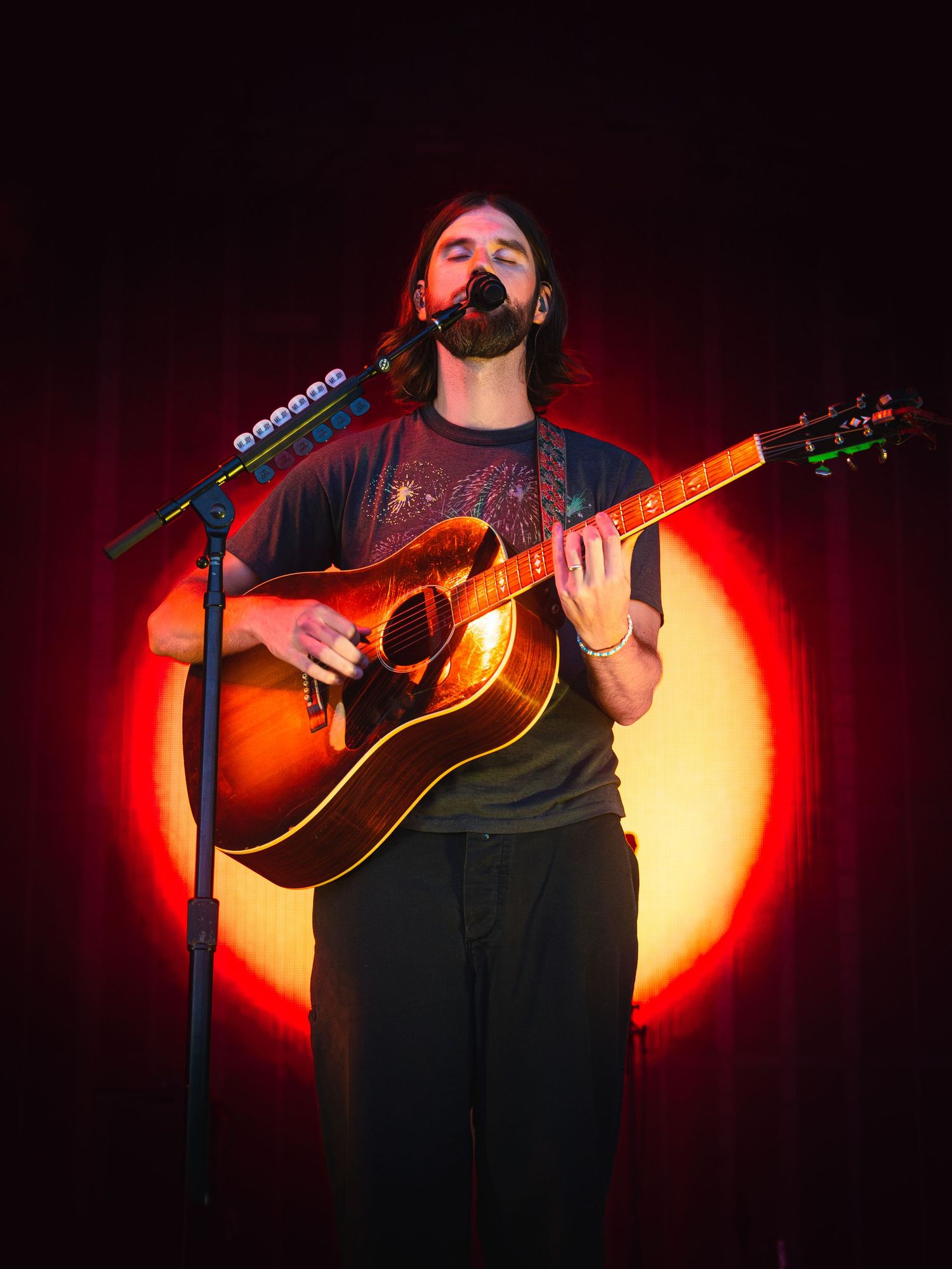 Male musician playing acoustic guitar on stage with dramatic red spotlight backdrop.