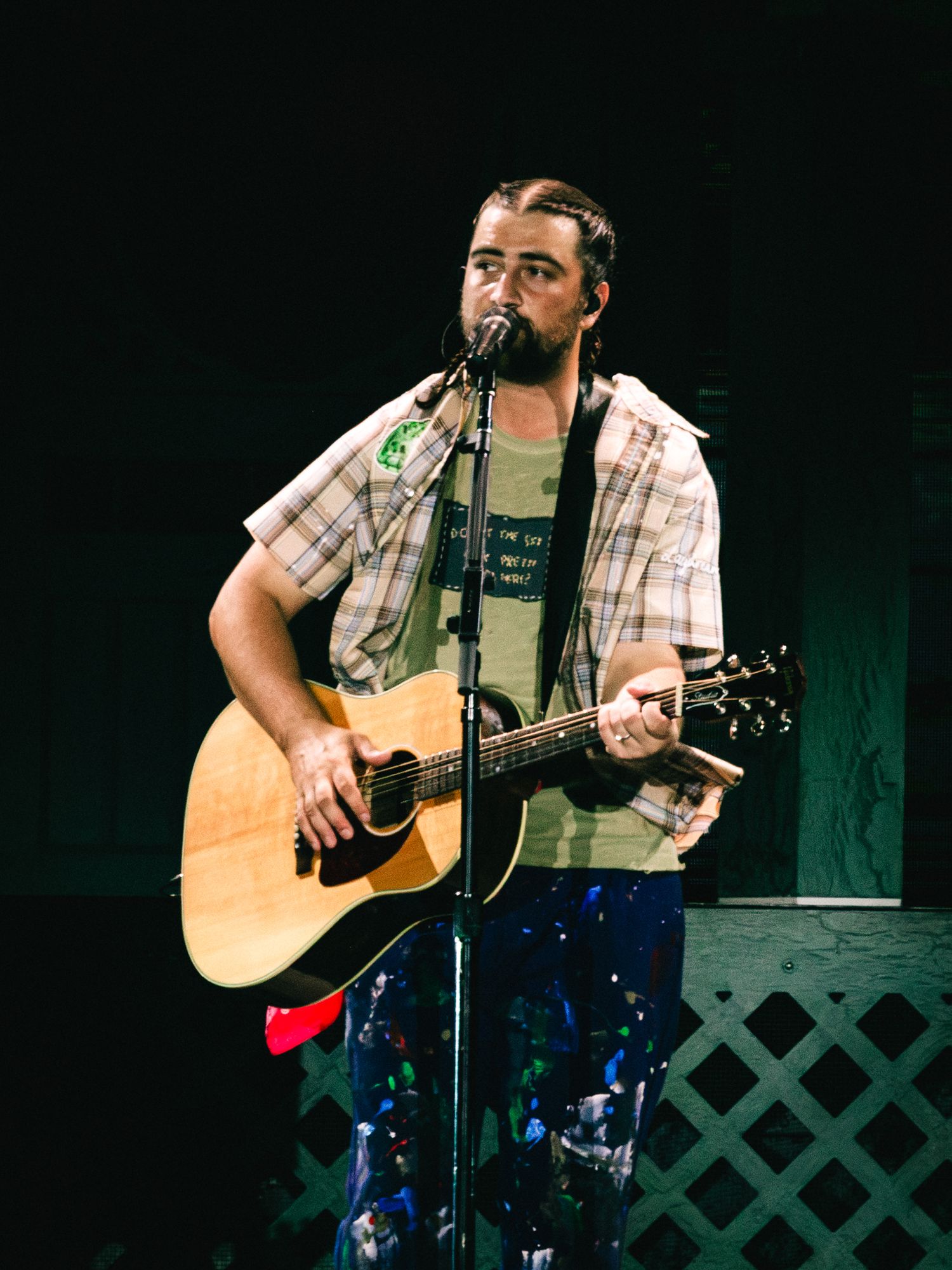 Man with braids playing acoustic guitar and singing into microphone on stage wearing plaid shirt.