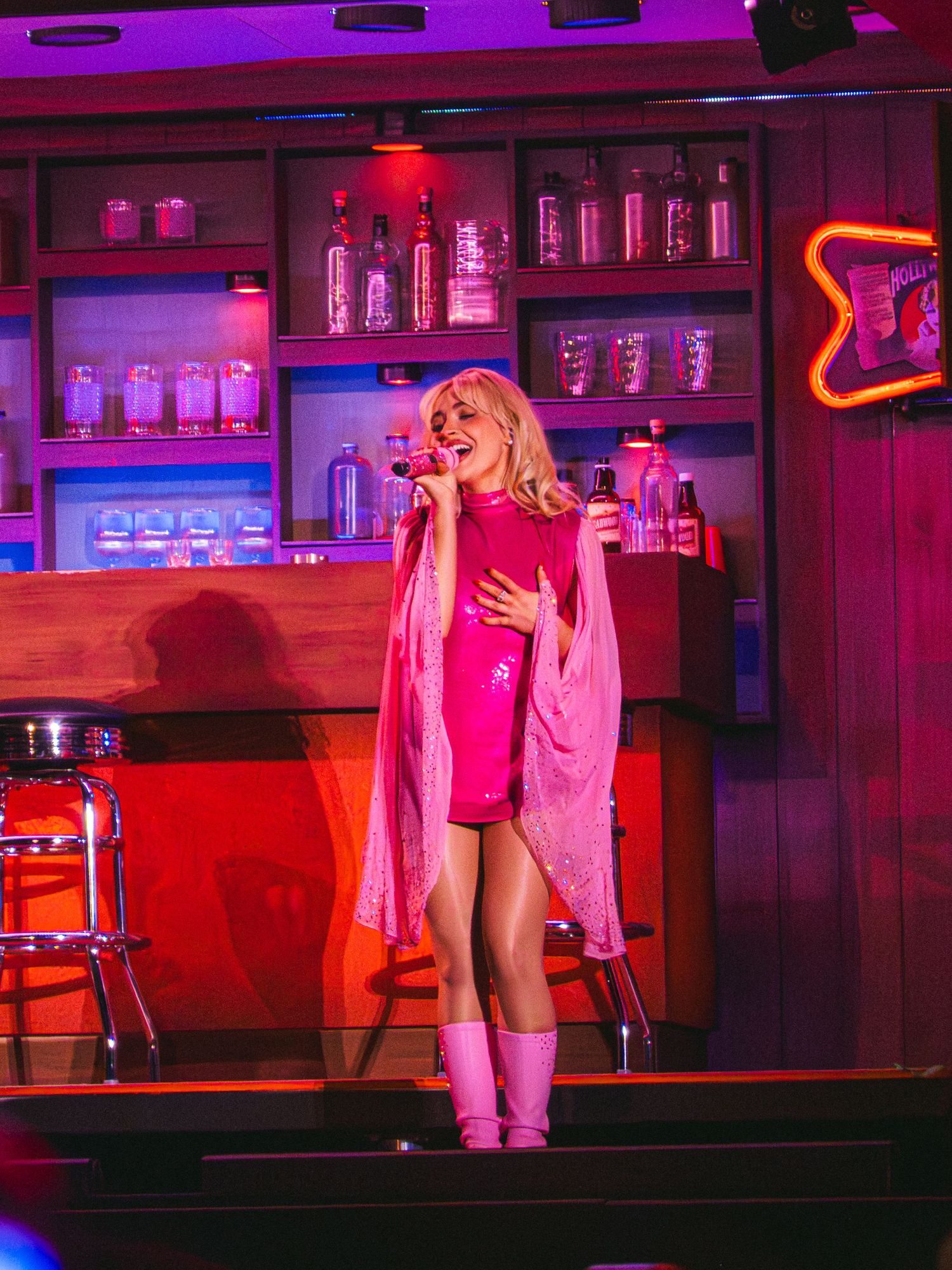 Performer in pink outfit singing on stage at a neon-lit bar set with bottles and pink lighting.