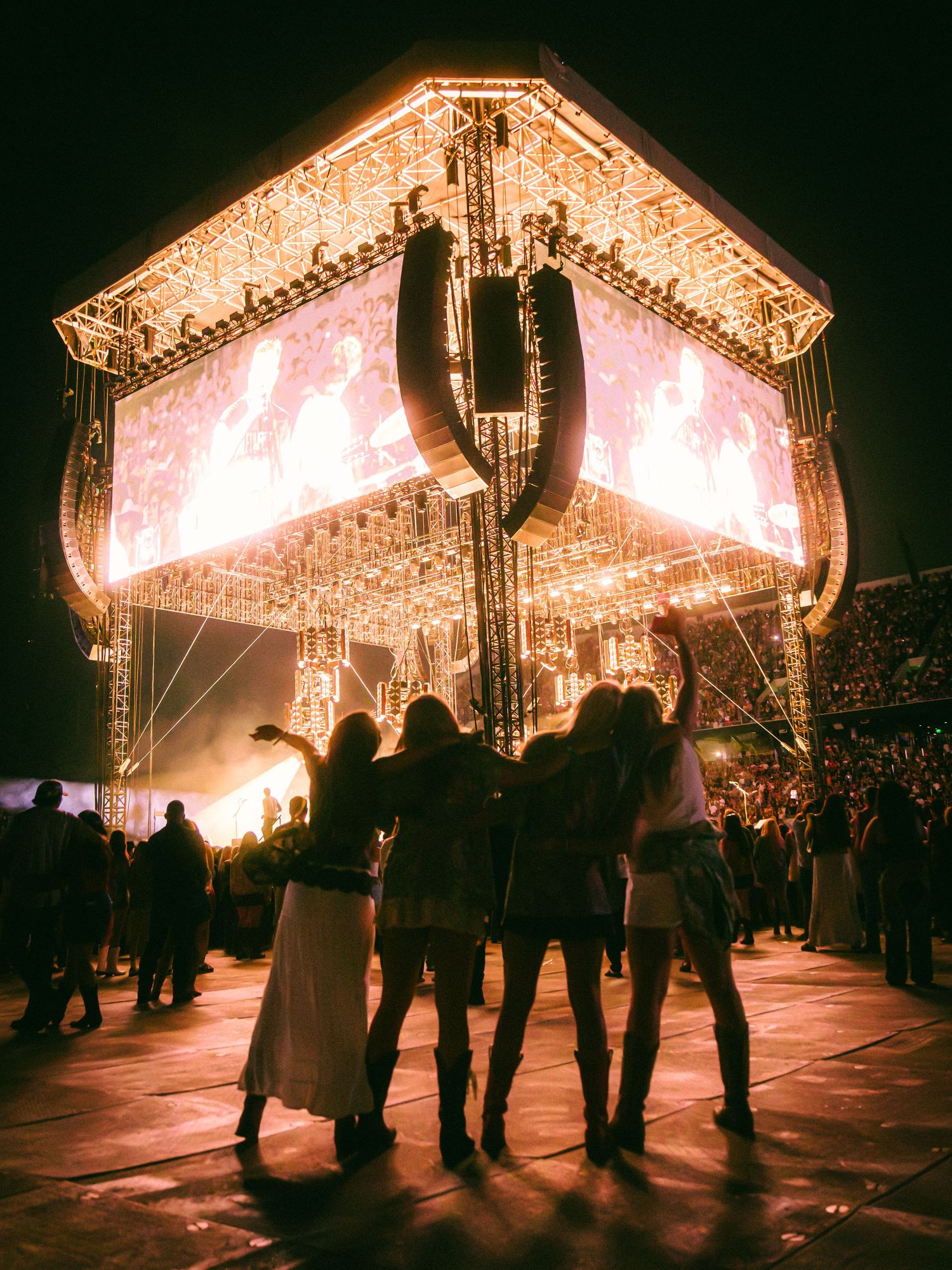Four women silhouetted against a glowing cube stage with LED lights at an outdoor music festival at night.