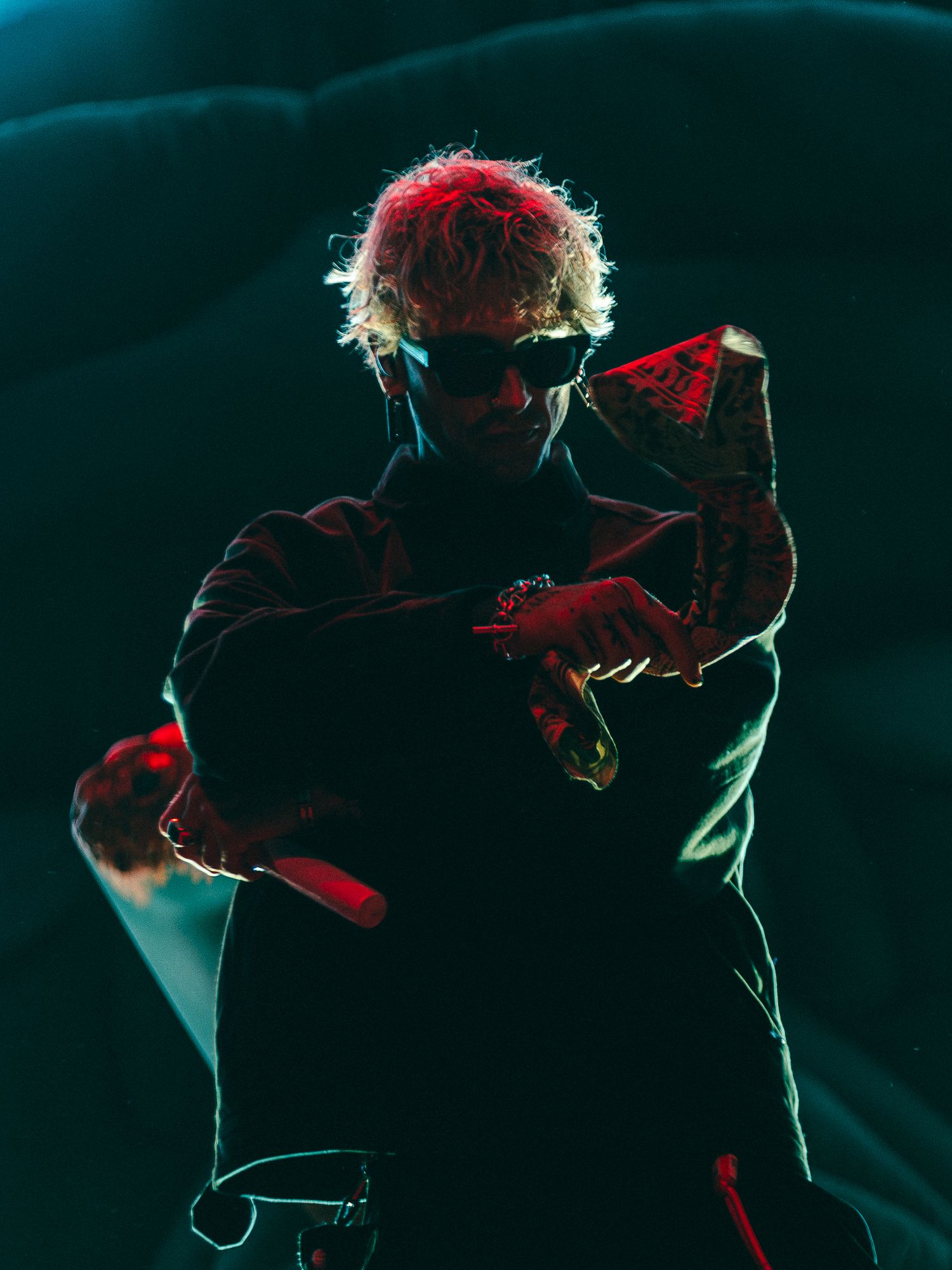 A performer with red hair and sunglasses poses dramatically on stage under teal and red lighting.