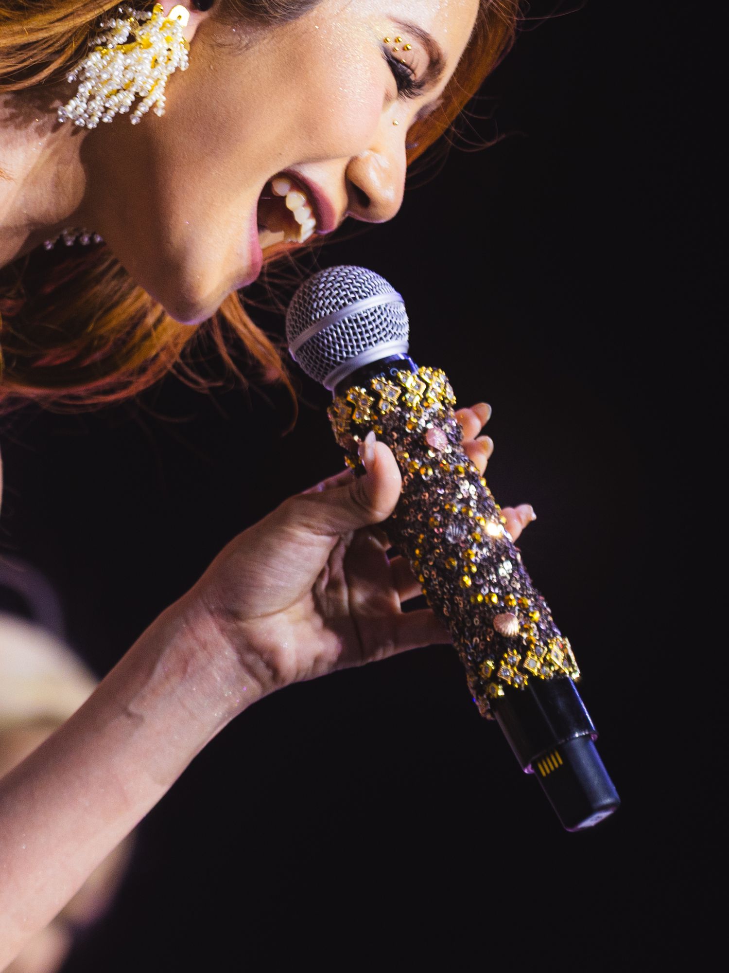 Woman singing passionately into a bejeweled microphone with gold and crystal embellishments on a dark stage.