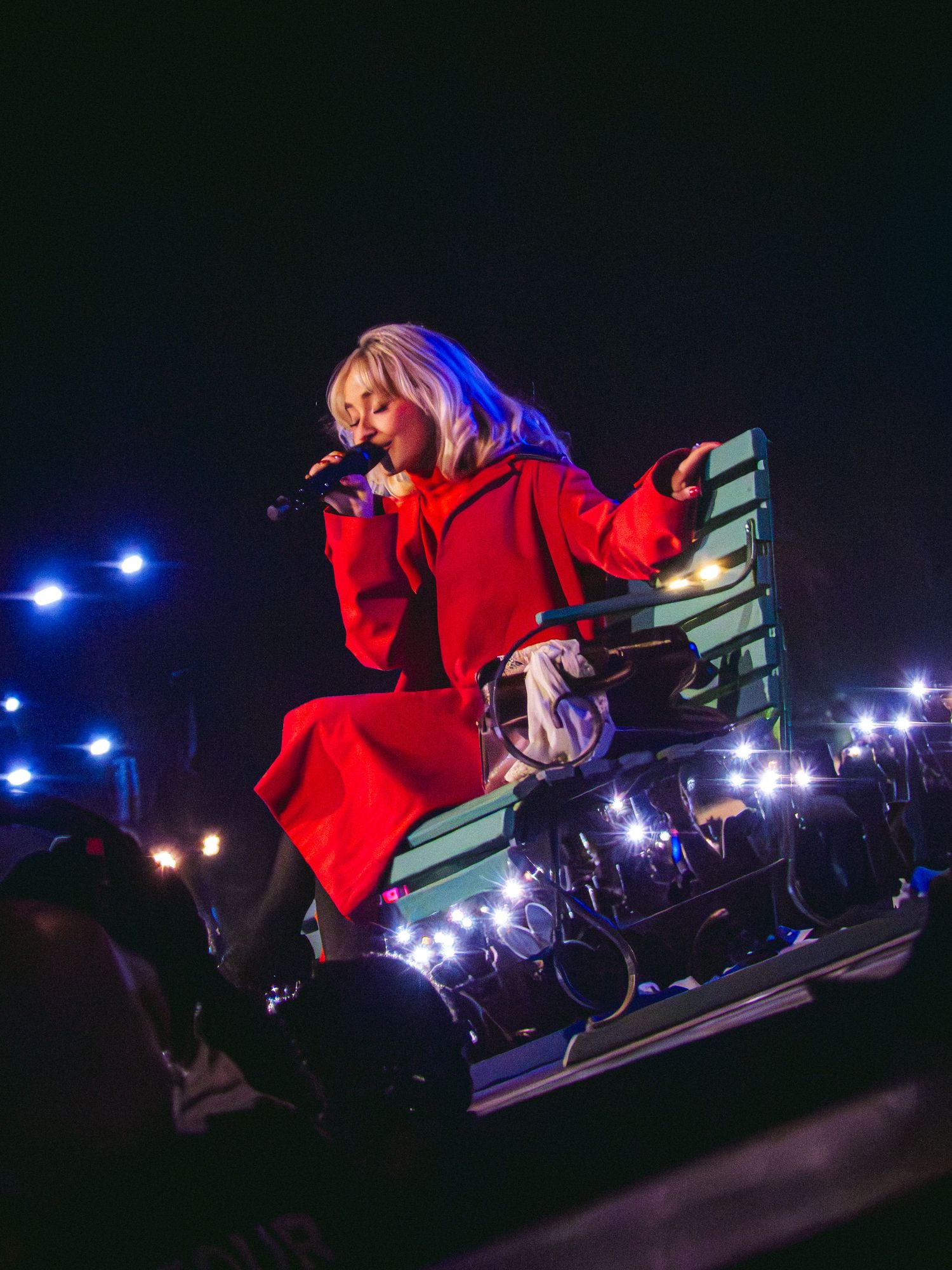 Female singer in red coat performs on stage sitting on a teal bench surrounded by bright lights at a concert.