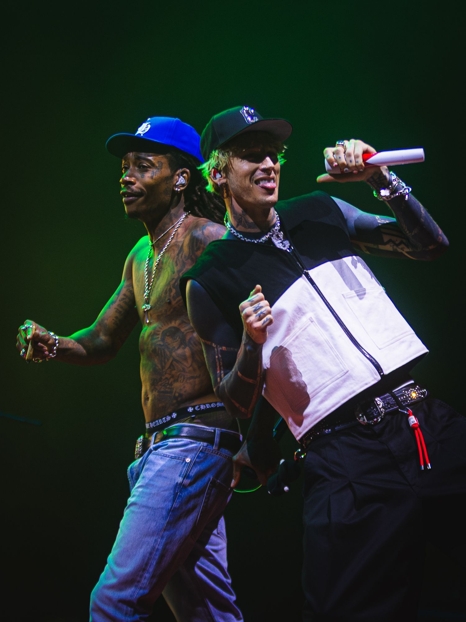Two tattooed male performers on stage, one shirtless in blue cap, other in black cap holding microphone under green lights.