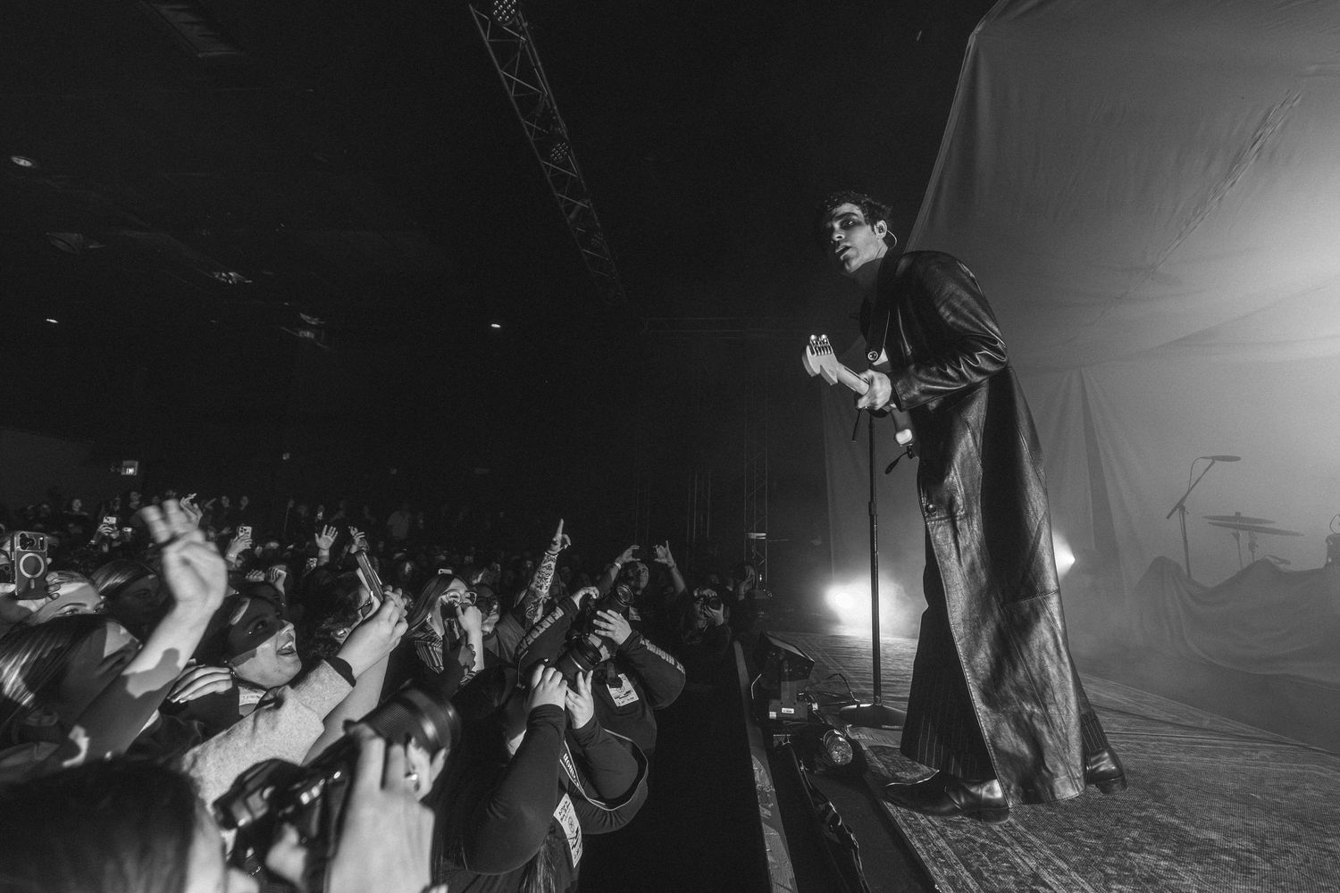 Concert crowd illuminated by stage lights raises their hands in excitement during a live performance at night.