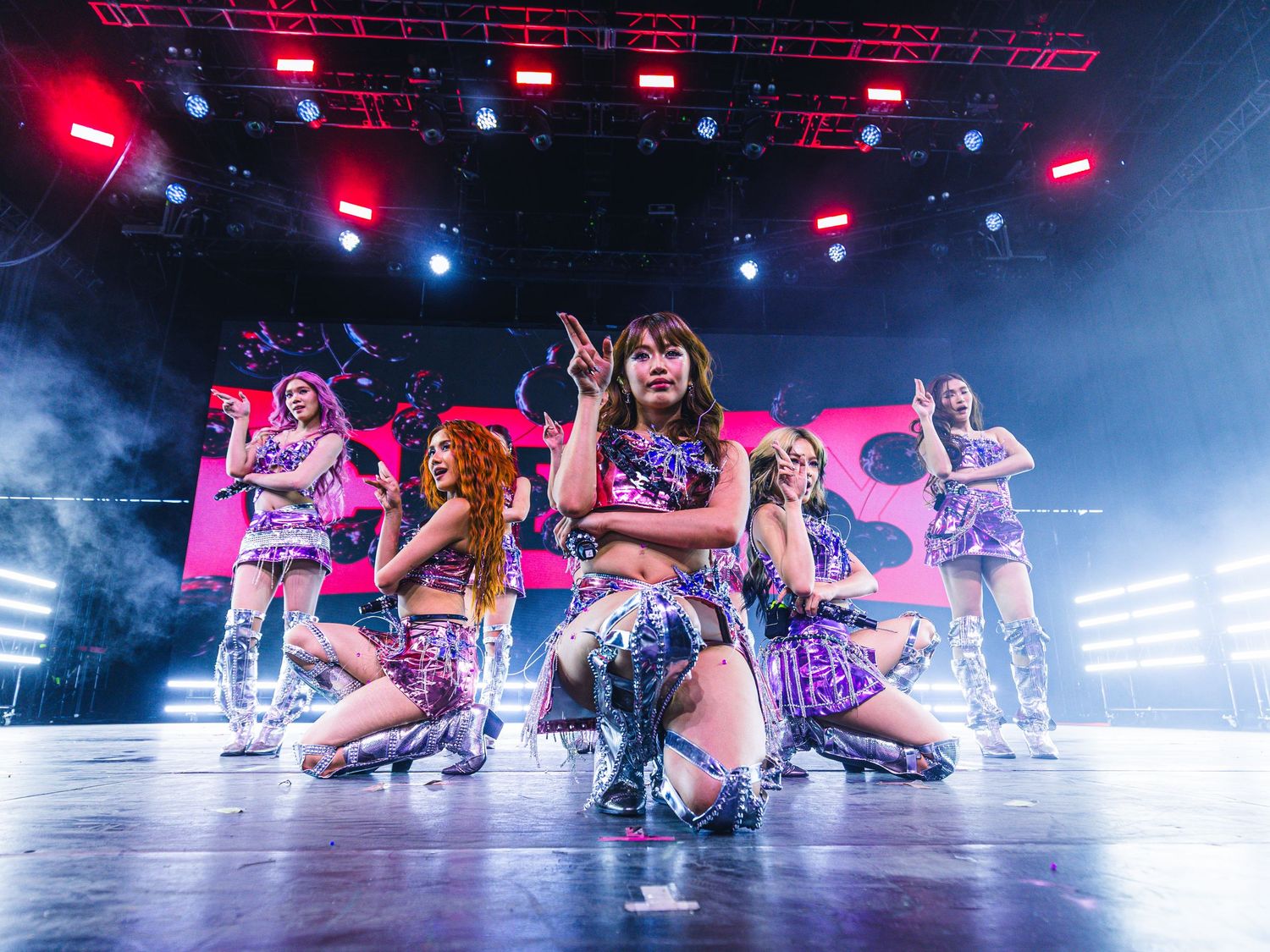 K-pop girl group performs energetically on a colorful stage with pink backdrop, smoke effects, and vibrant purple costumes.