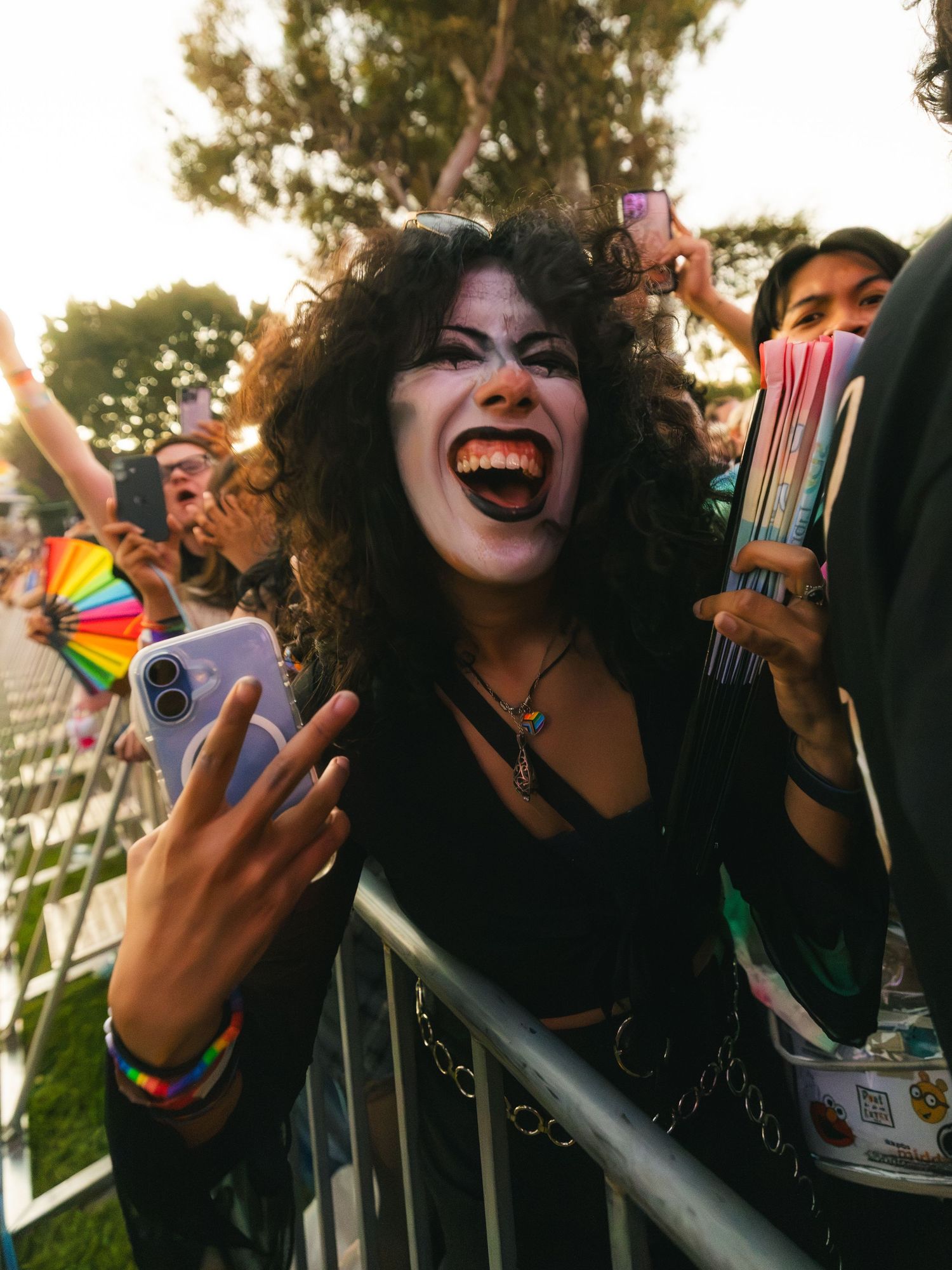Excited festival-goer with curly hair and vampire makeup makes rock sign at outdoor music concert.