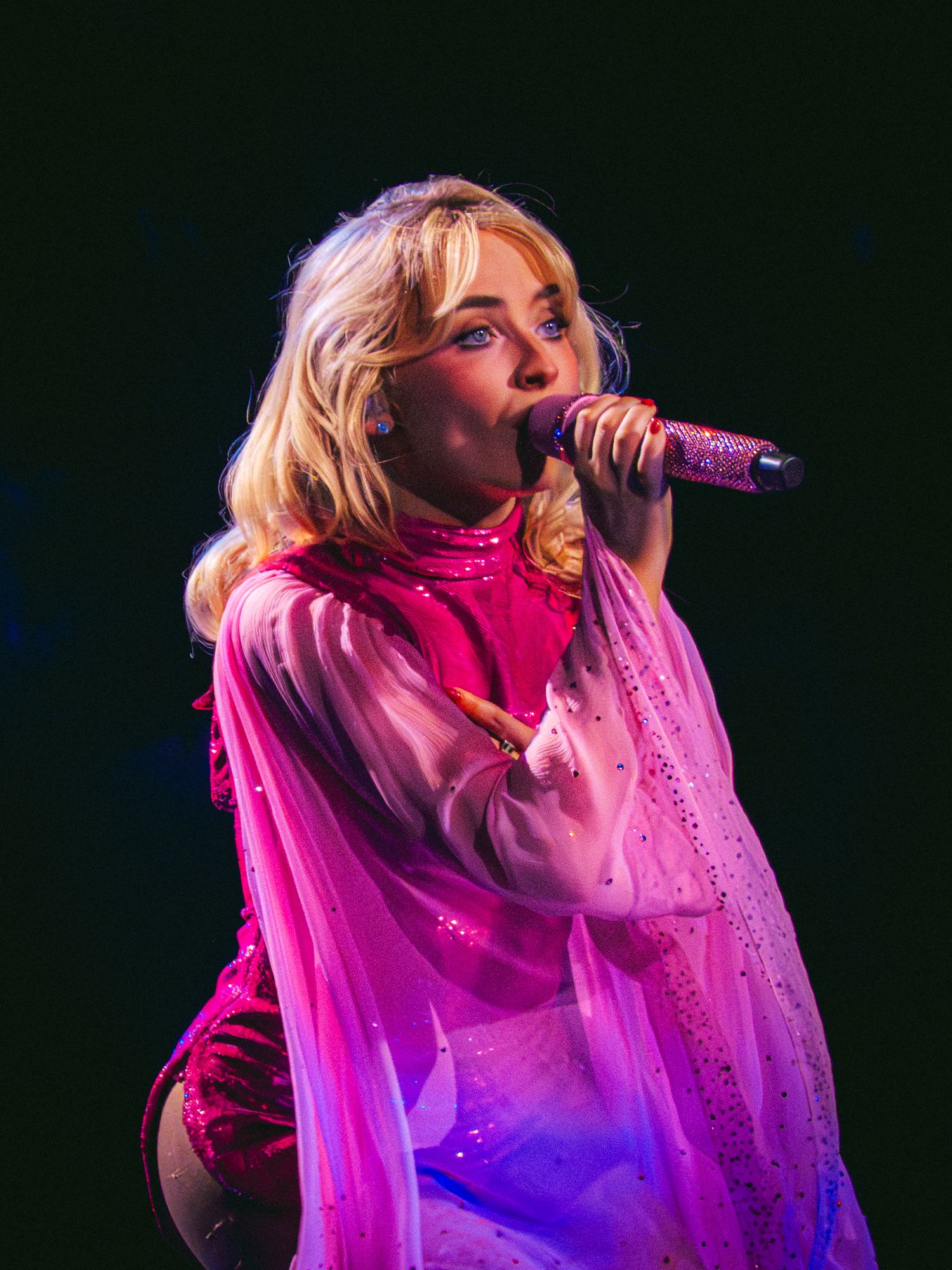 Female pop singer performing on stage in pink outfit, holding glittery microphone under dramatic lighting.