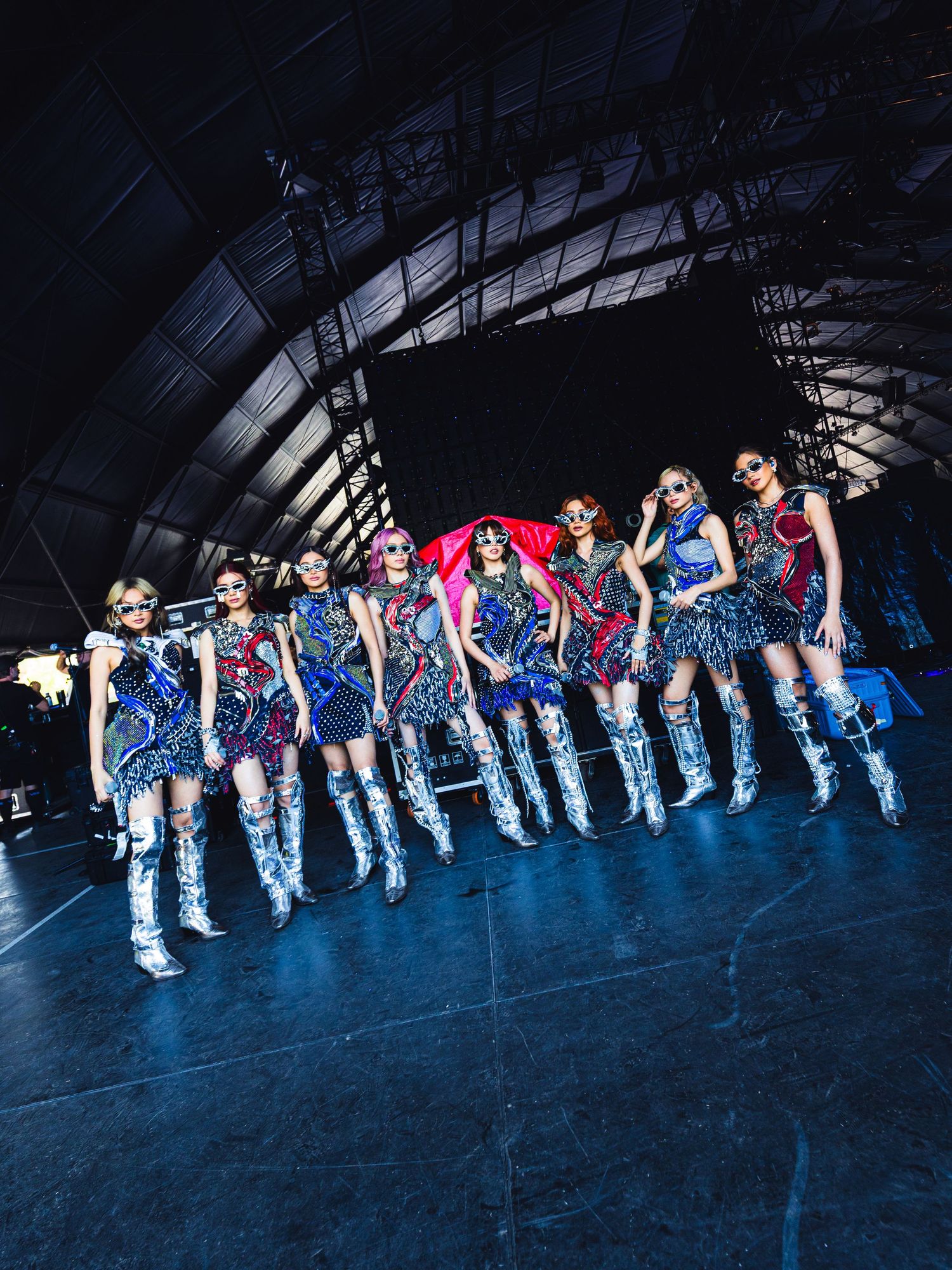 A group of women in futuristic metallic outfits and silver boots pose on a stage under dramatic lighting.