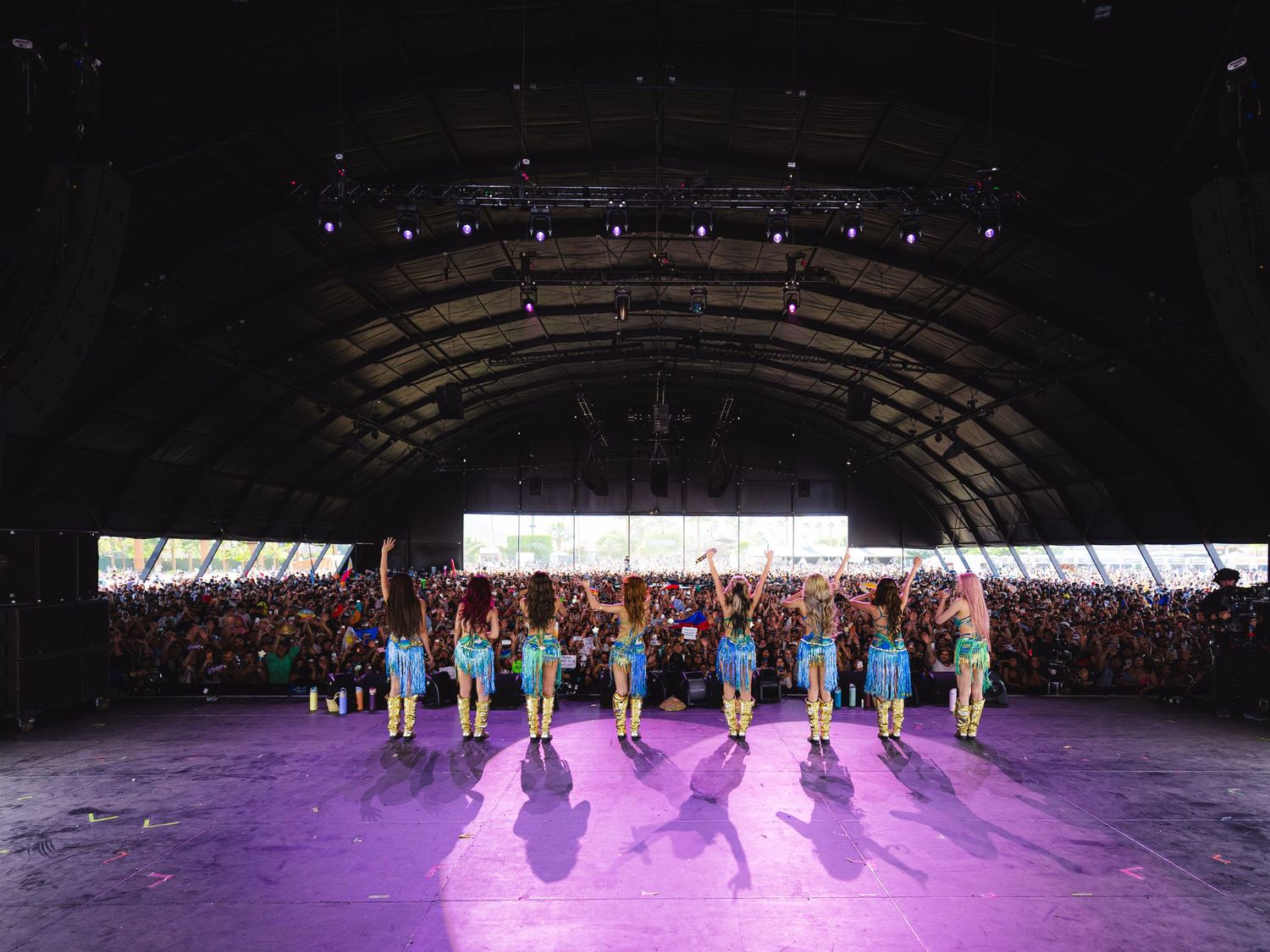 Performers in blue costumes on a purple-lit stage under an arched venue roof before a large festival crowd.