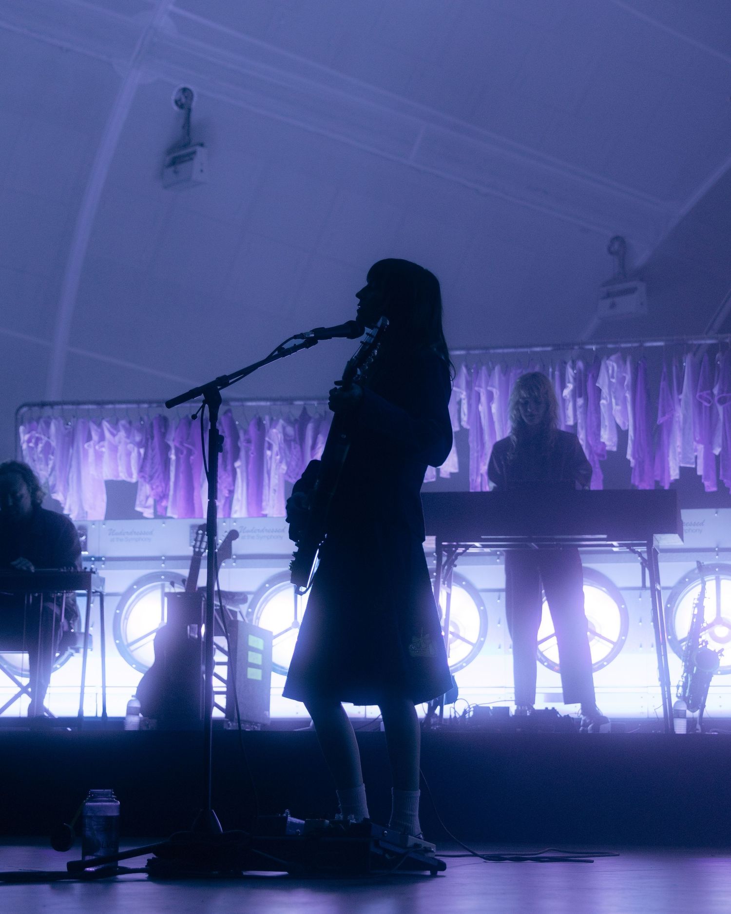 Silhouetted performer on stage with purple backlighting and band members visible behind illuminated backdrop.