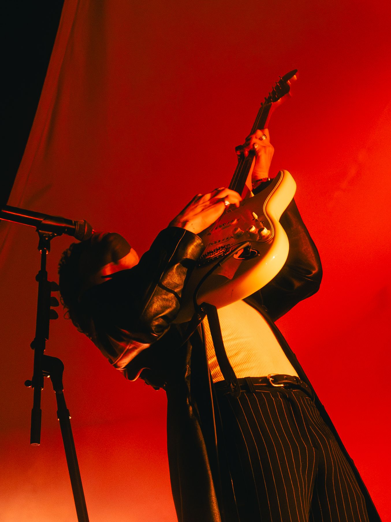 Performer in black and white striped outfit leaning back dramatically against microphone stand under red stage lighting.