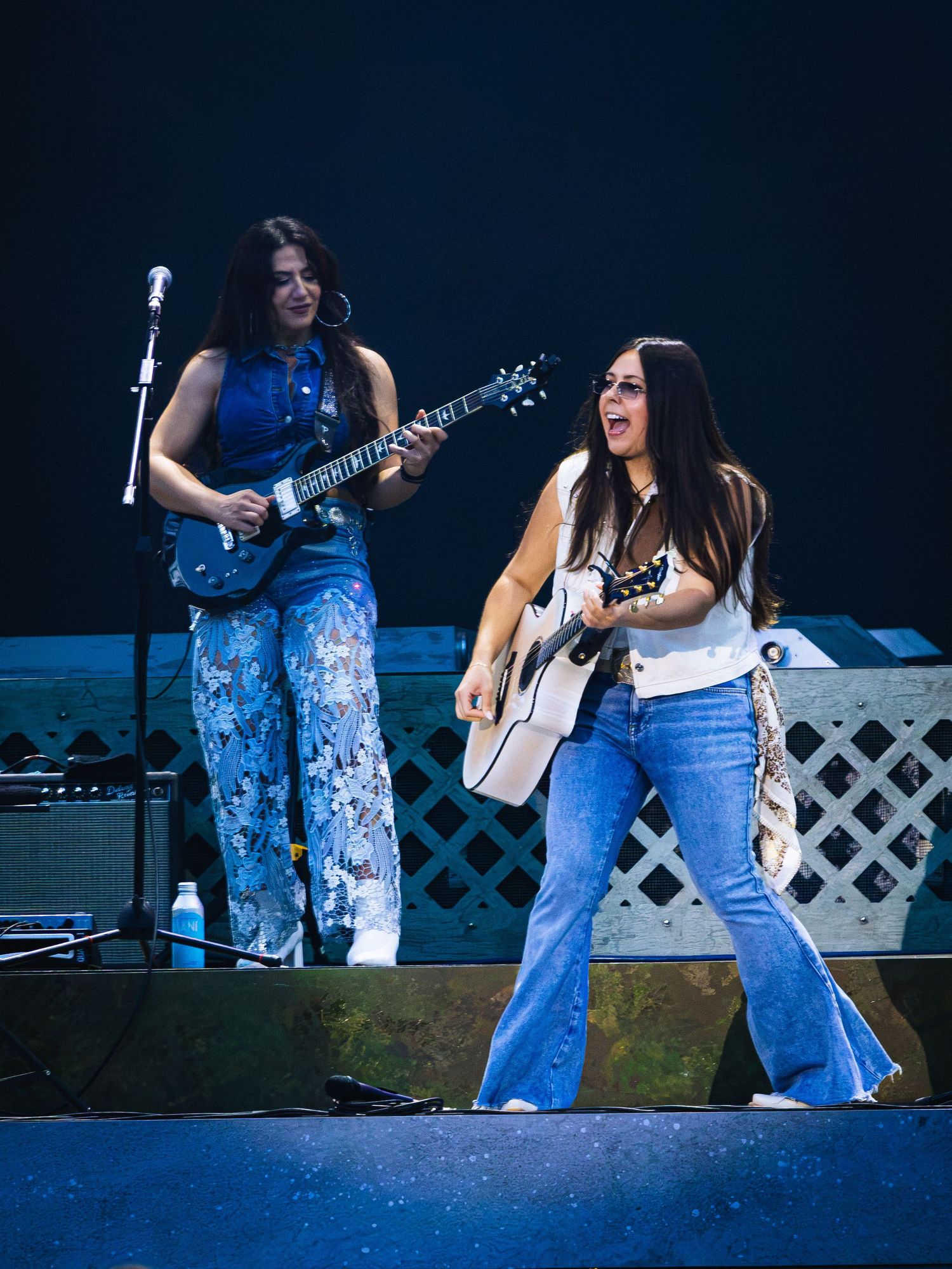 Two women performing on stage with guitars, wearing jeans, smiling under blue stage lights.