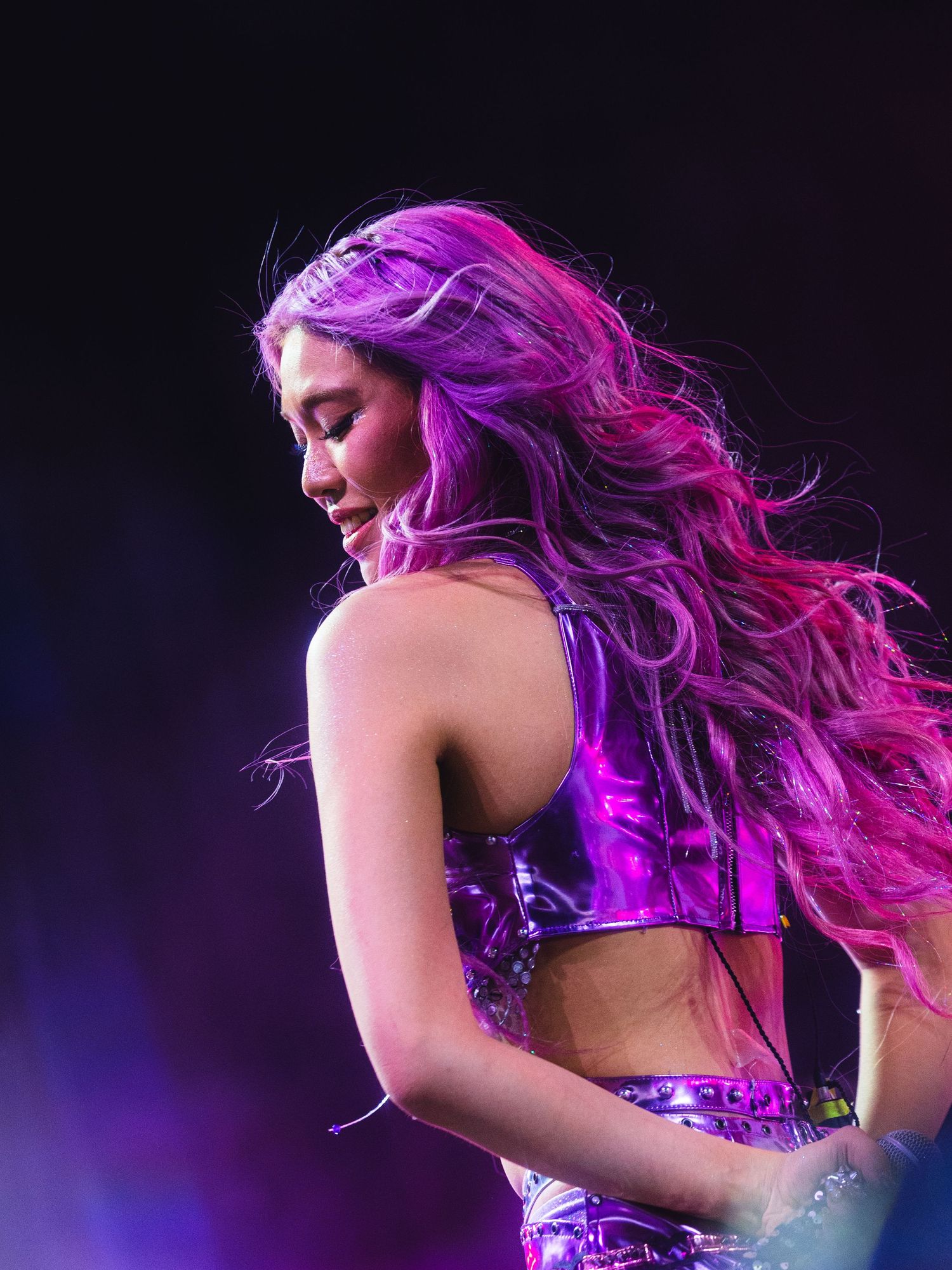 A performer with long purple-pink hair wearing a metallic crop top dances on stage under vibrant purple stage lighting.