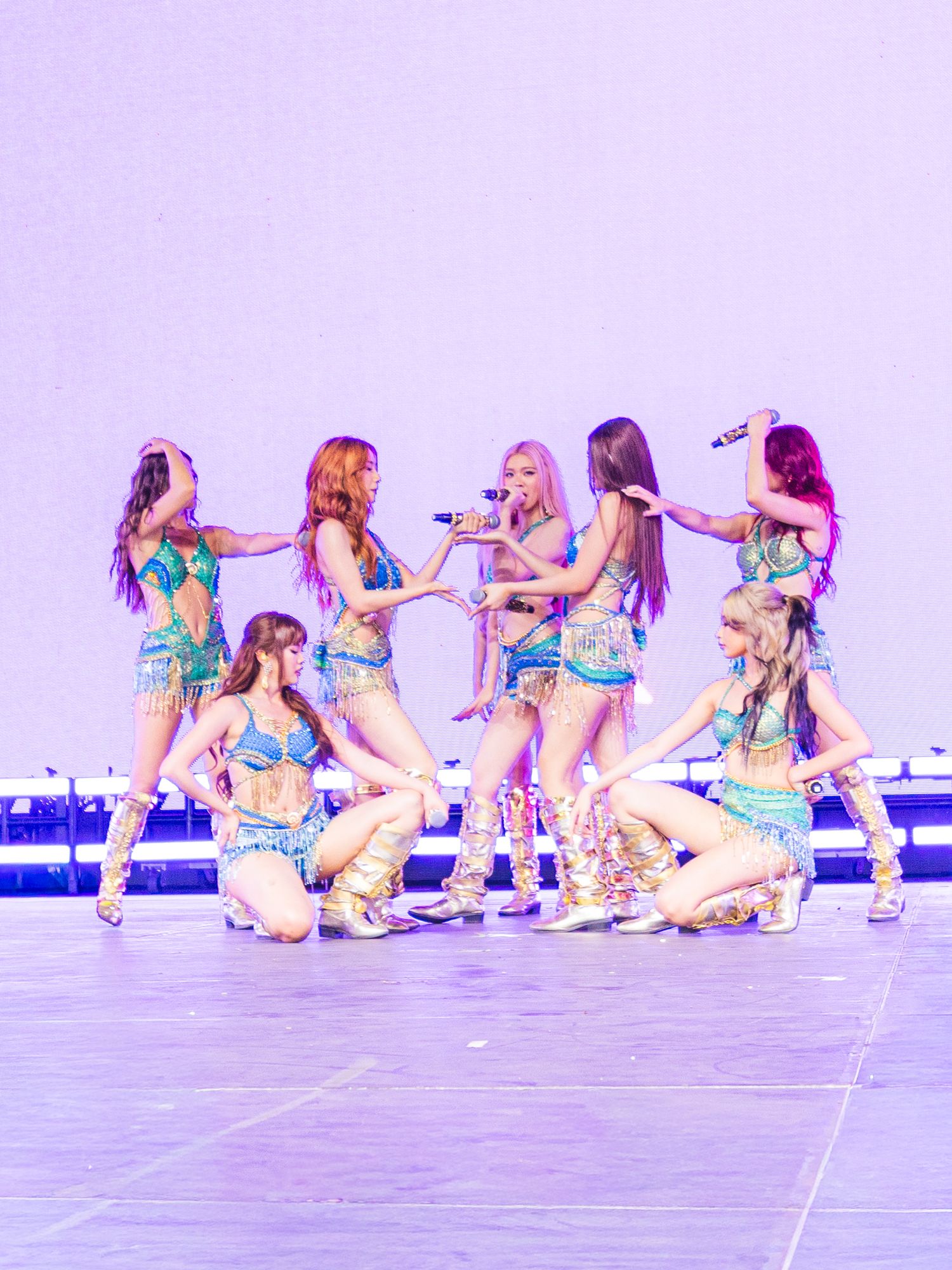 K-pop girl group performs on stage in matching iridescent teal outfits, dancing in formation under purple stage lighting.