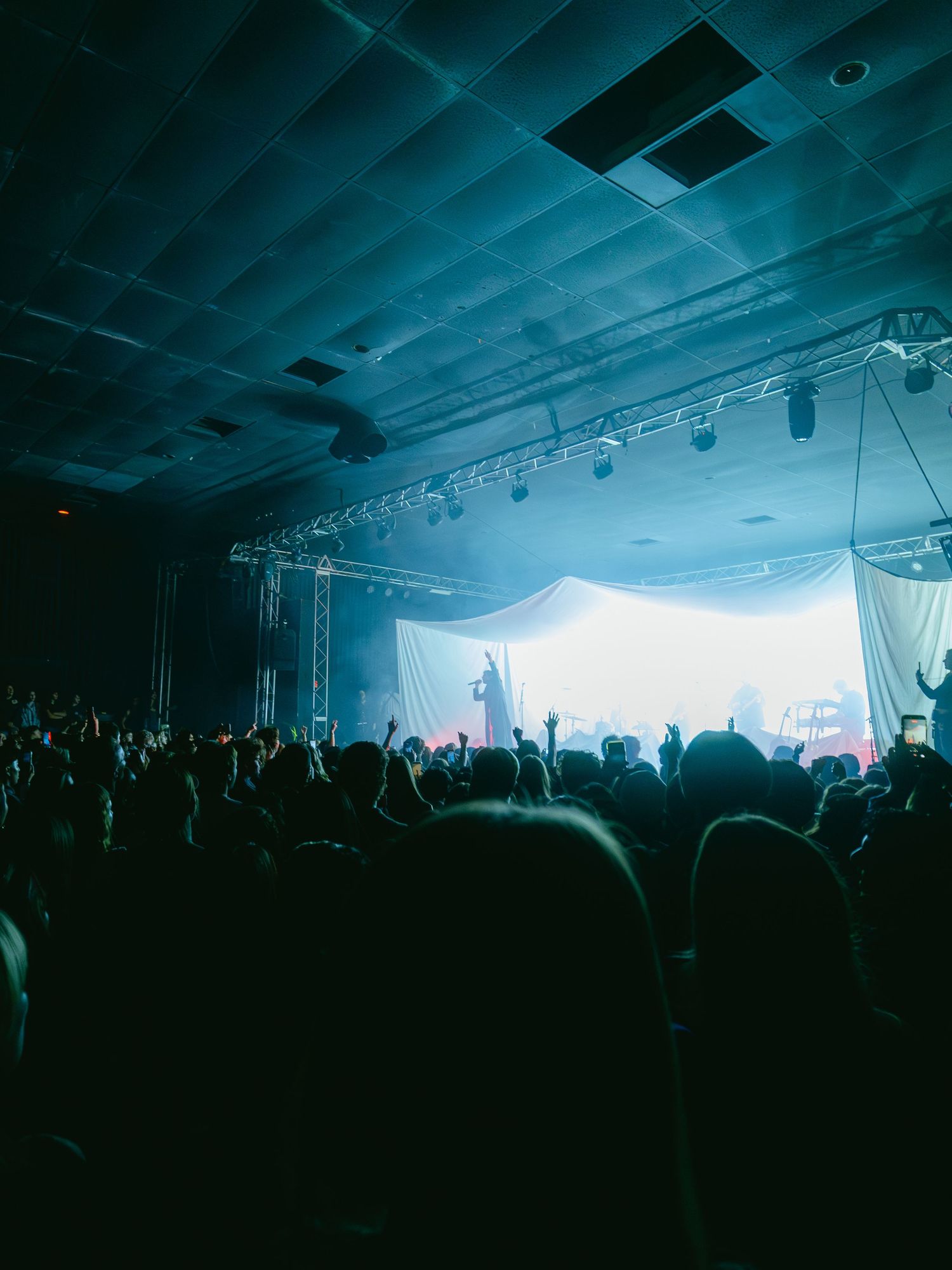 Silhouetted crowd at a concert venue under moody blue lighting and hazy atmosphere during a live performance.