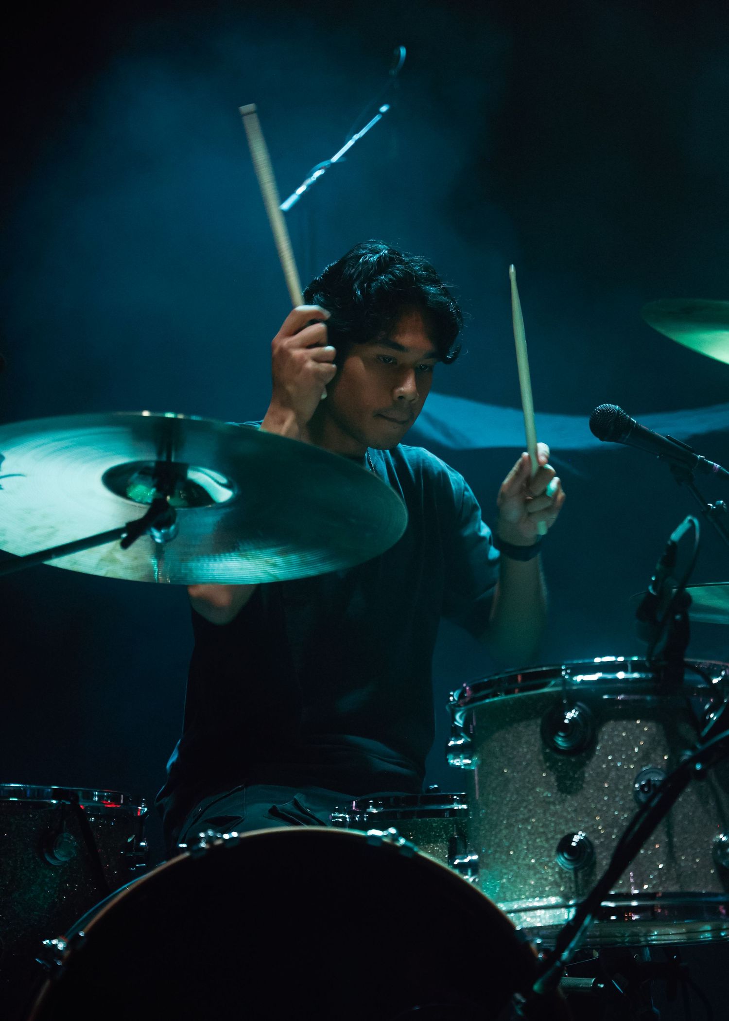 Drummer performing intensely behind drum kit with blue stage lighting and cymbals.