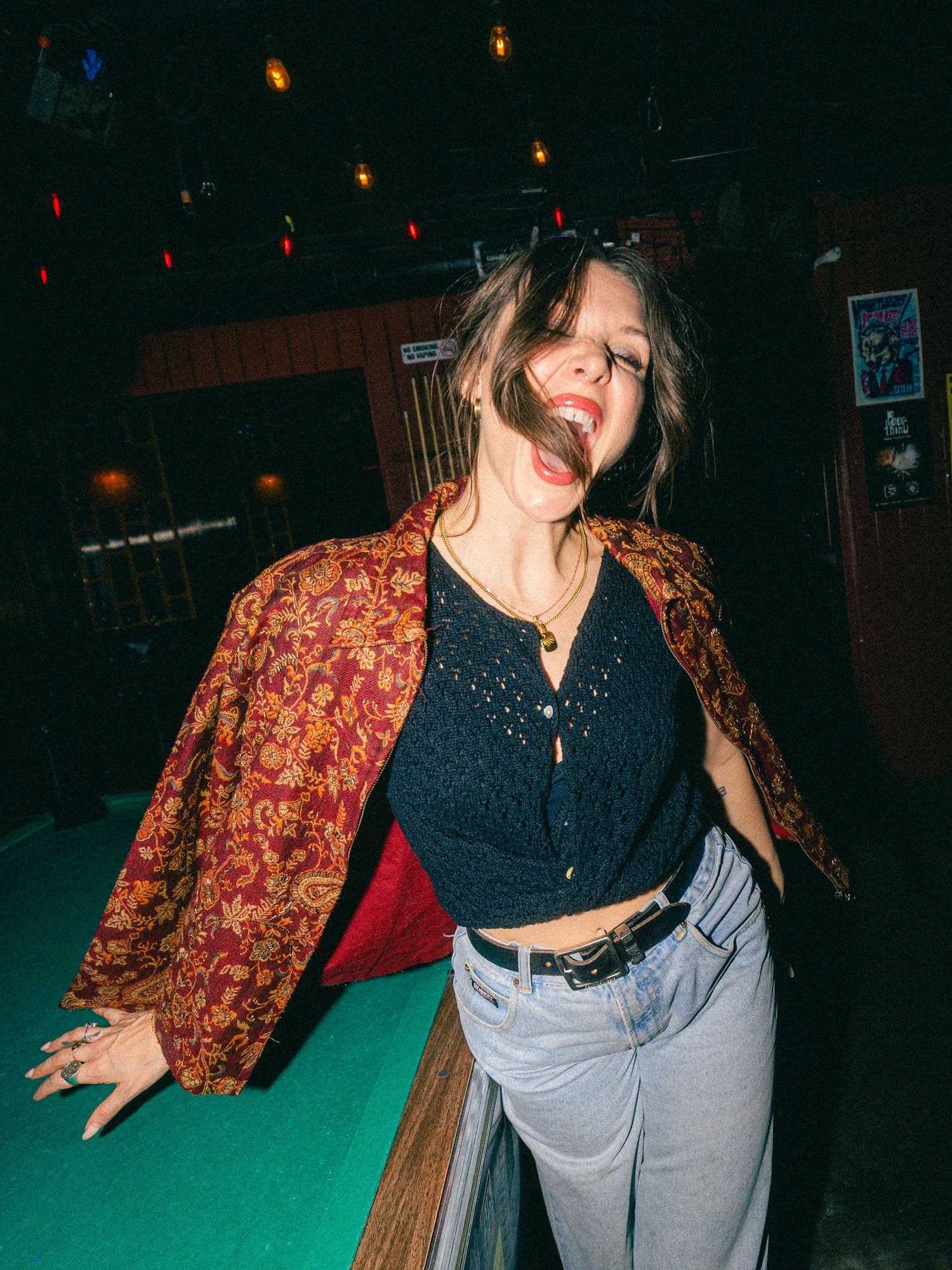 Woman in floral kimono jacket and black crop top poses playfully near a pool table in a dimly lit bar.