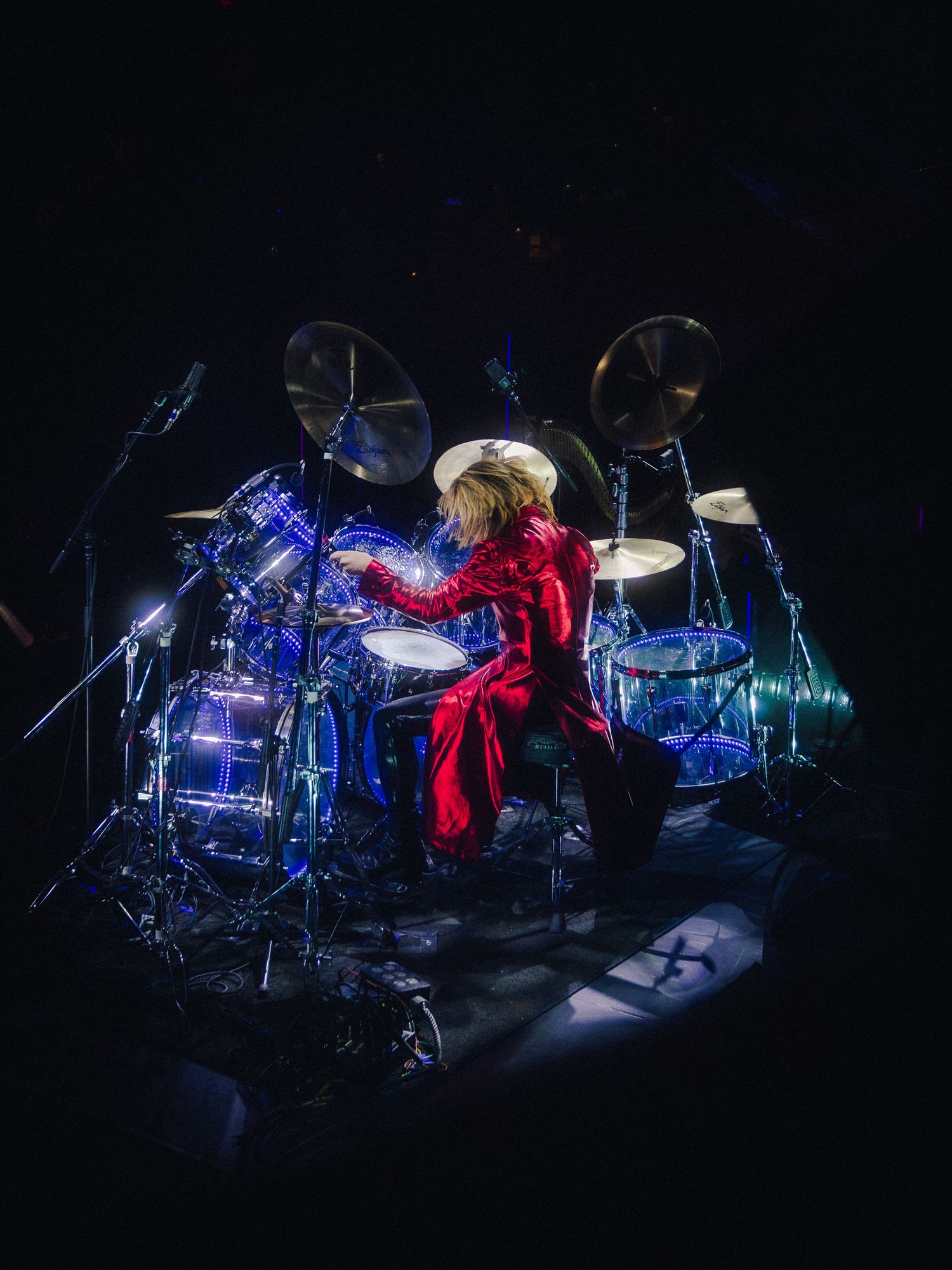 Drummer in red suit performing on illuminated blue drum kit on dark concert stage.