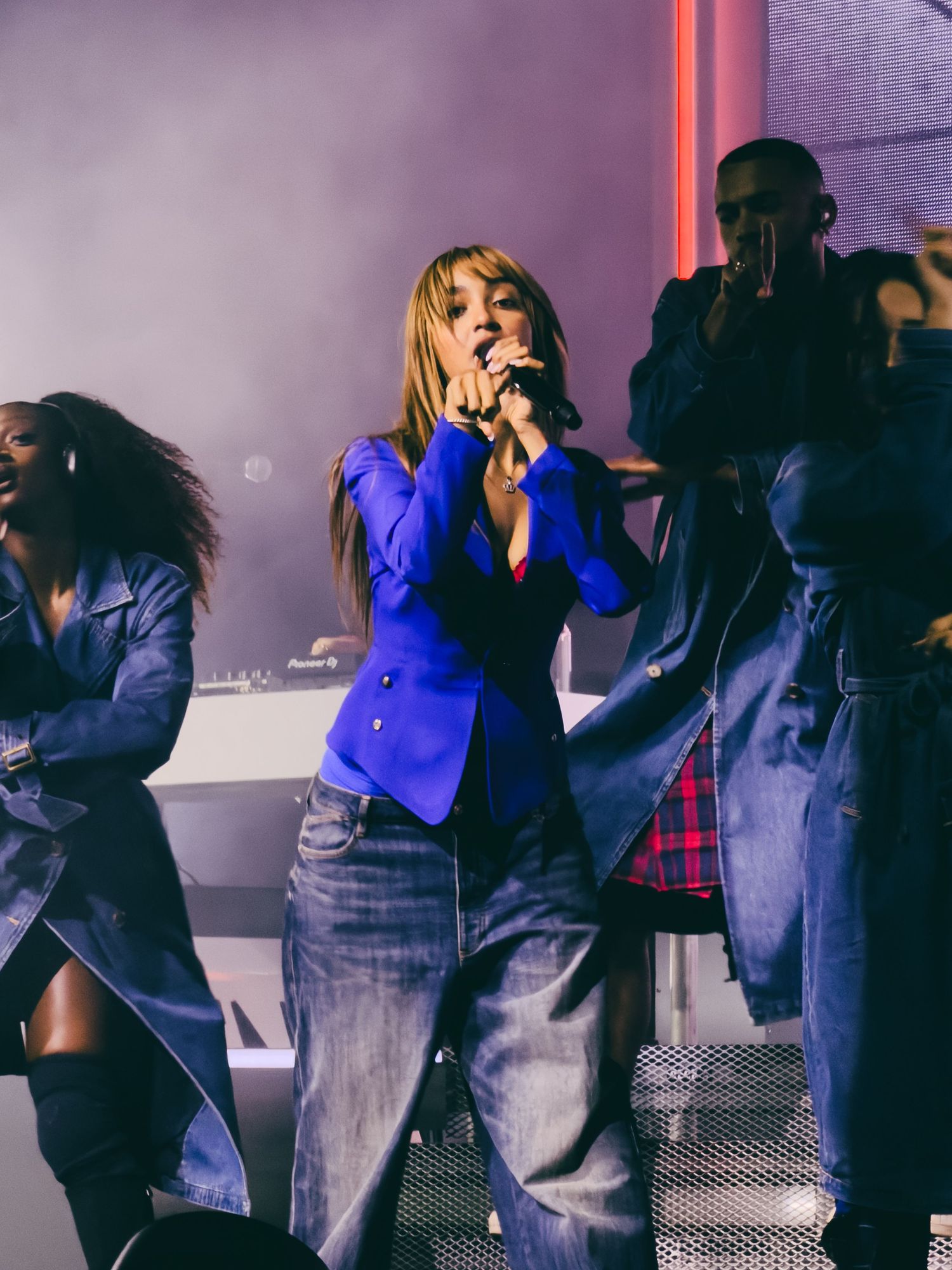 A woman in a blue blazer and jeans performs on stage with backup dancers and a DJ in a smoky venue.