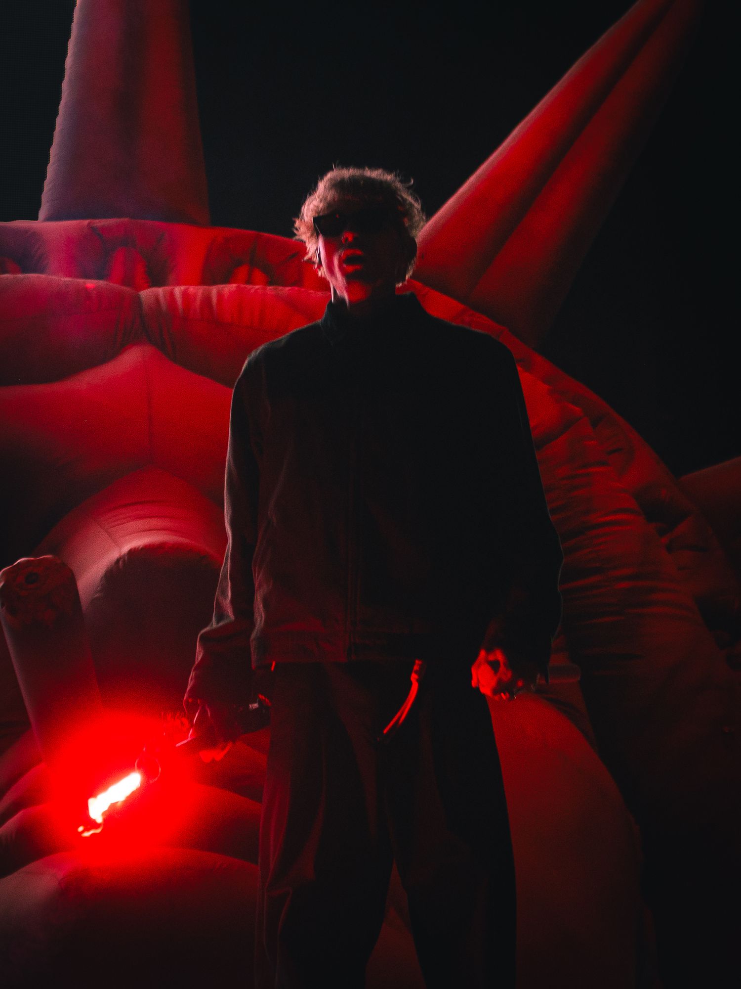 Silhouetted figure stands before red inflatable structures holding a glowing red light in dramatic dark atmosphere.