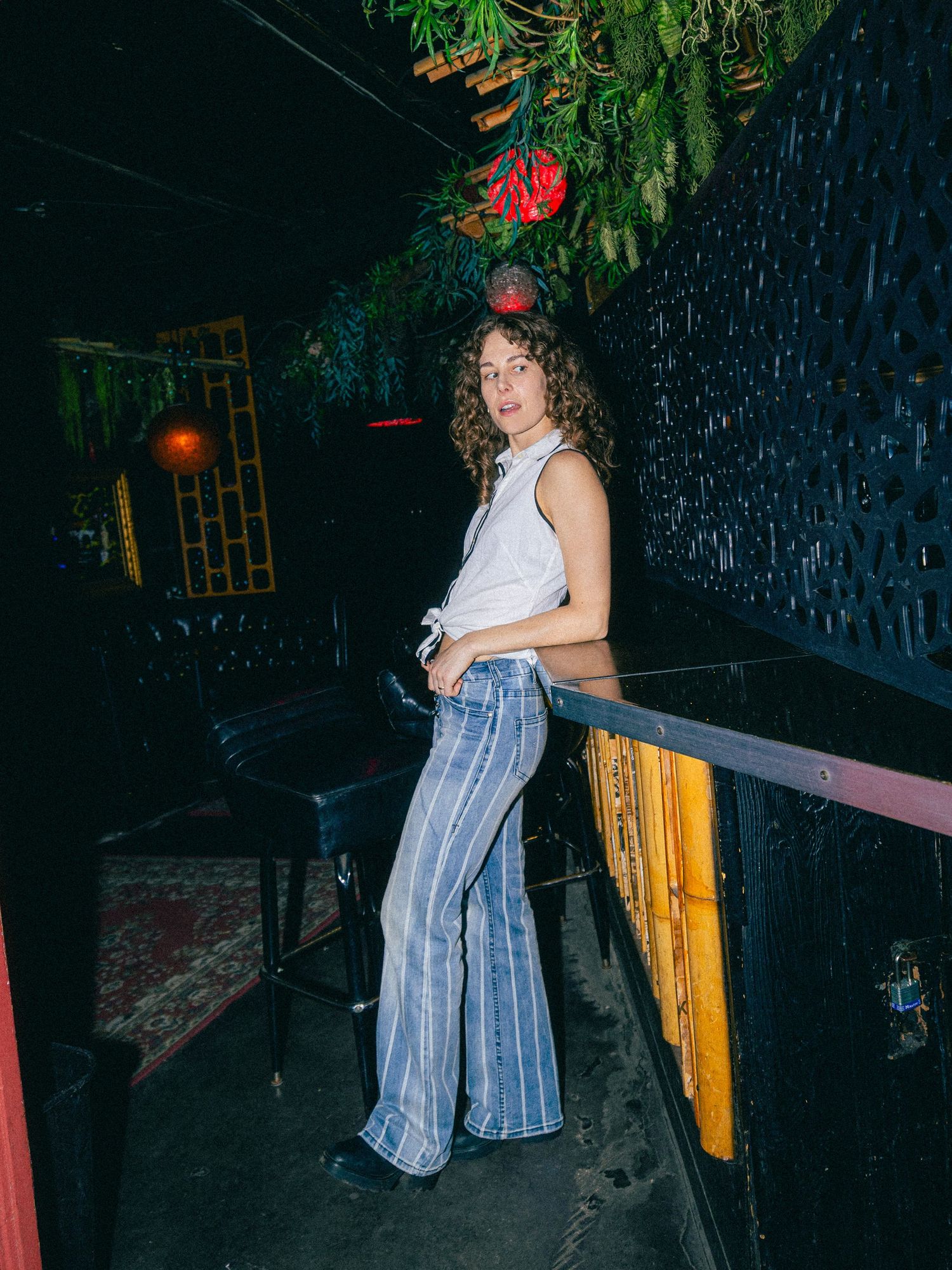 Woman in striped flare jeans and white sleeveless top posing in a dimly lit bar with lush greenery.