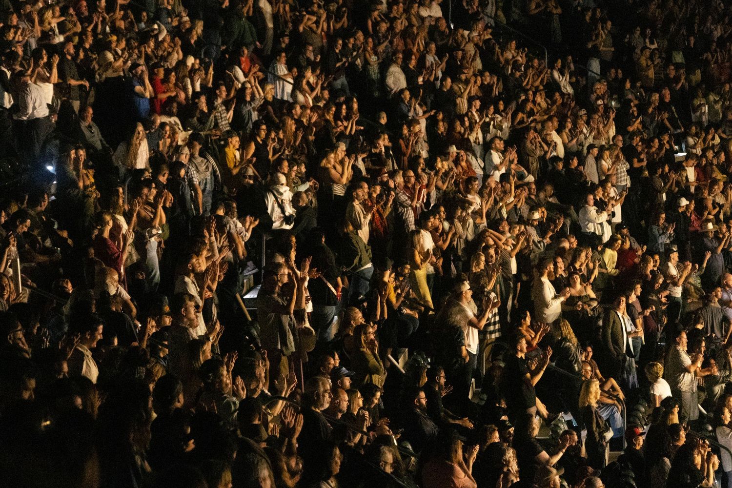 Massive crowd of concert attendees filling a dark venue with hands raised during a live performance.