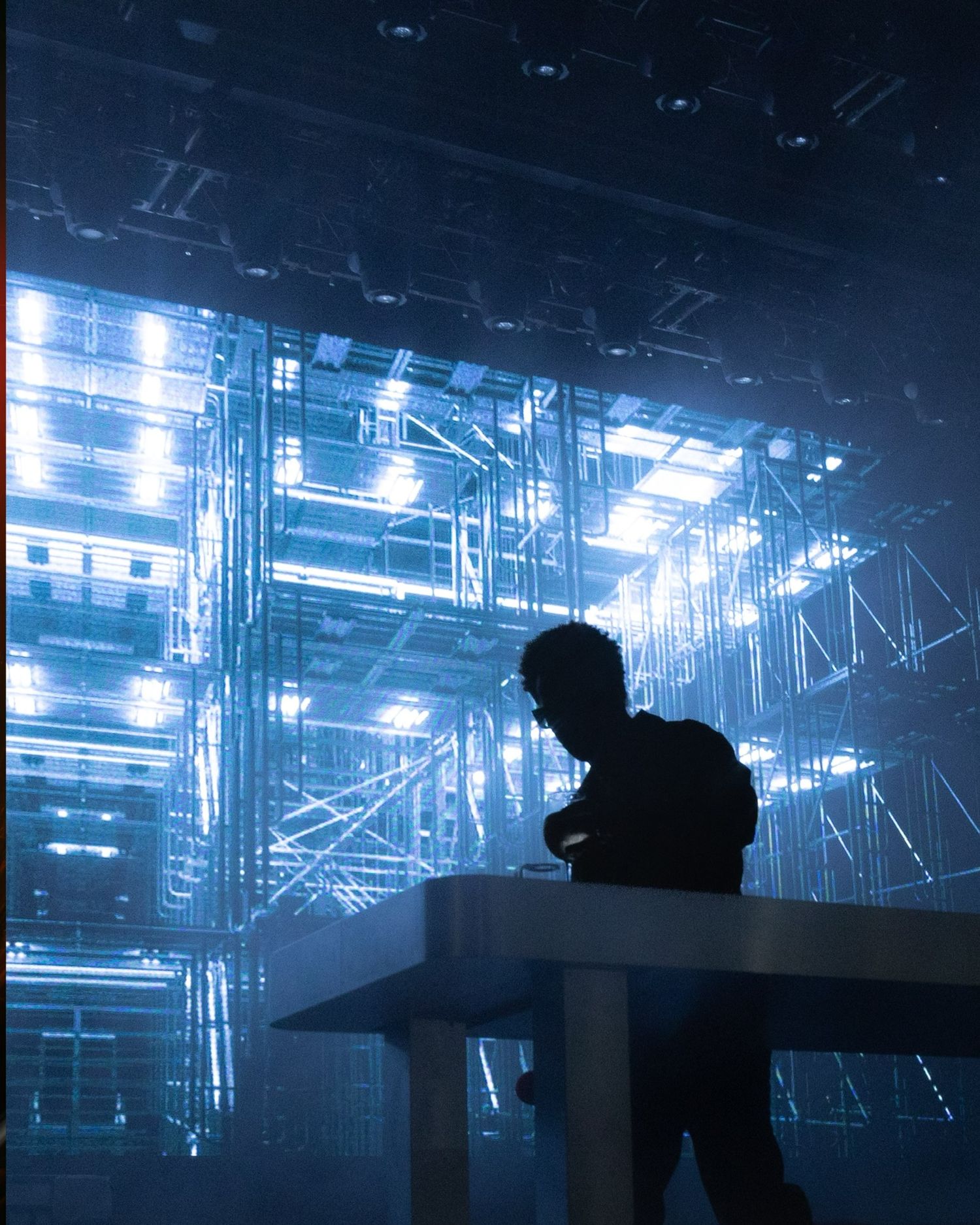 Silhouetted figures behind DJ equipment with blue stage lighting and fog effects.