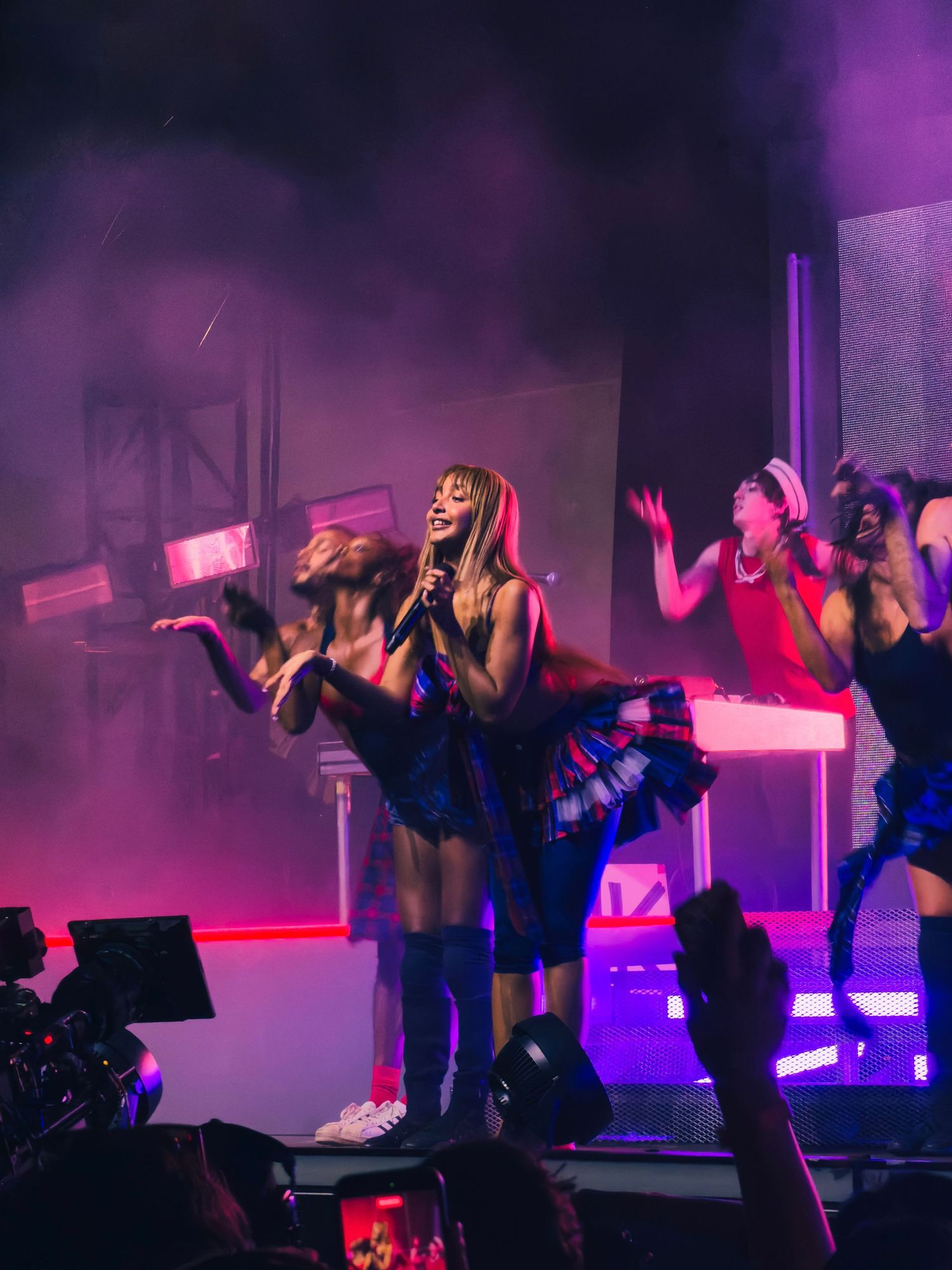 Pop singer performs with backup dancers in colorful outfits under purple stage lighting at concert.