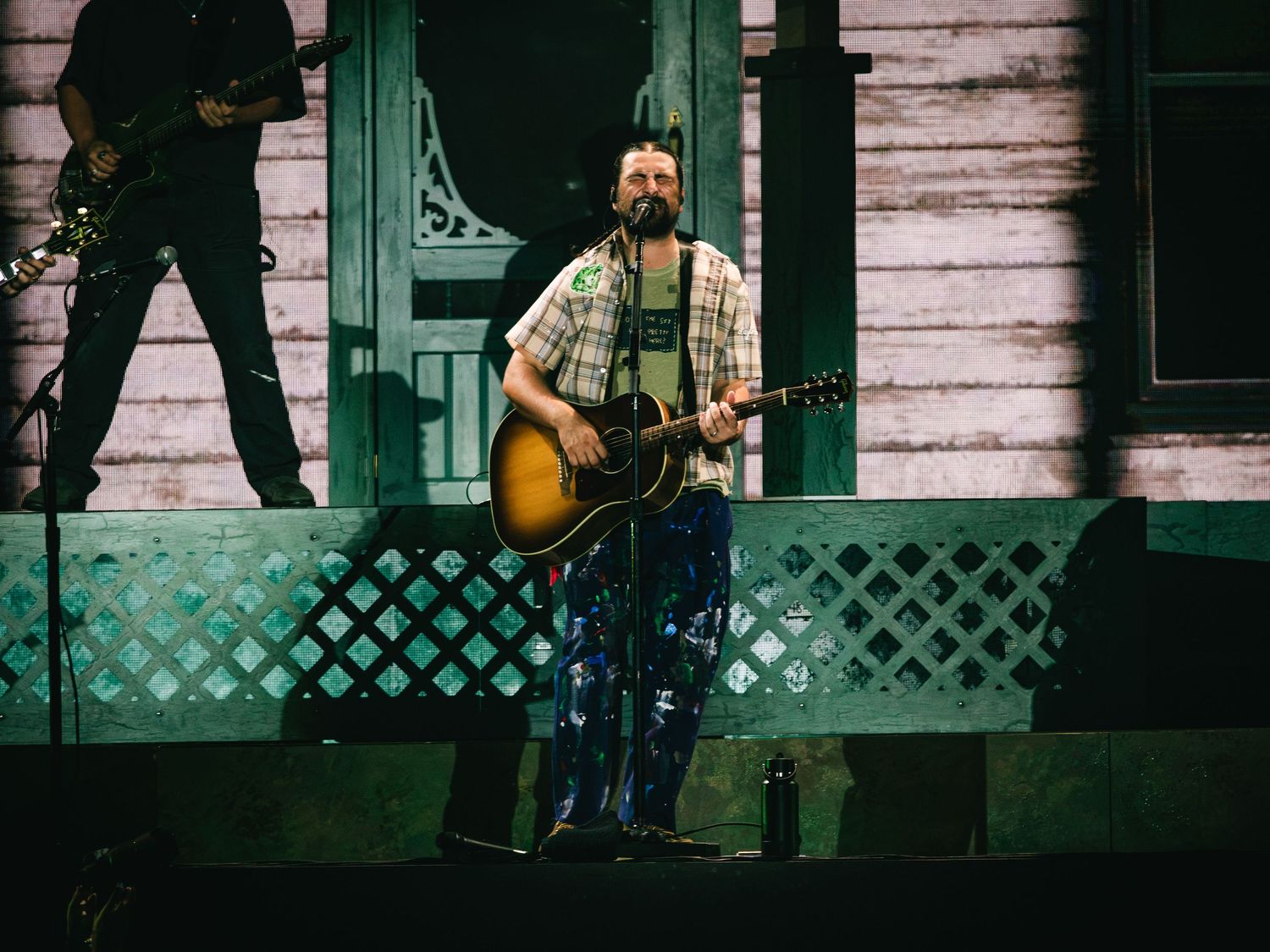 Musician performs on stage with acoustic guitar, wearing plaid shirt, in front of a rustic house backdrop.