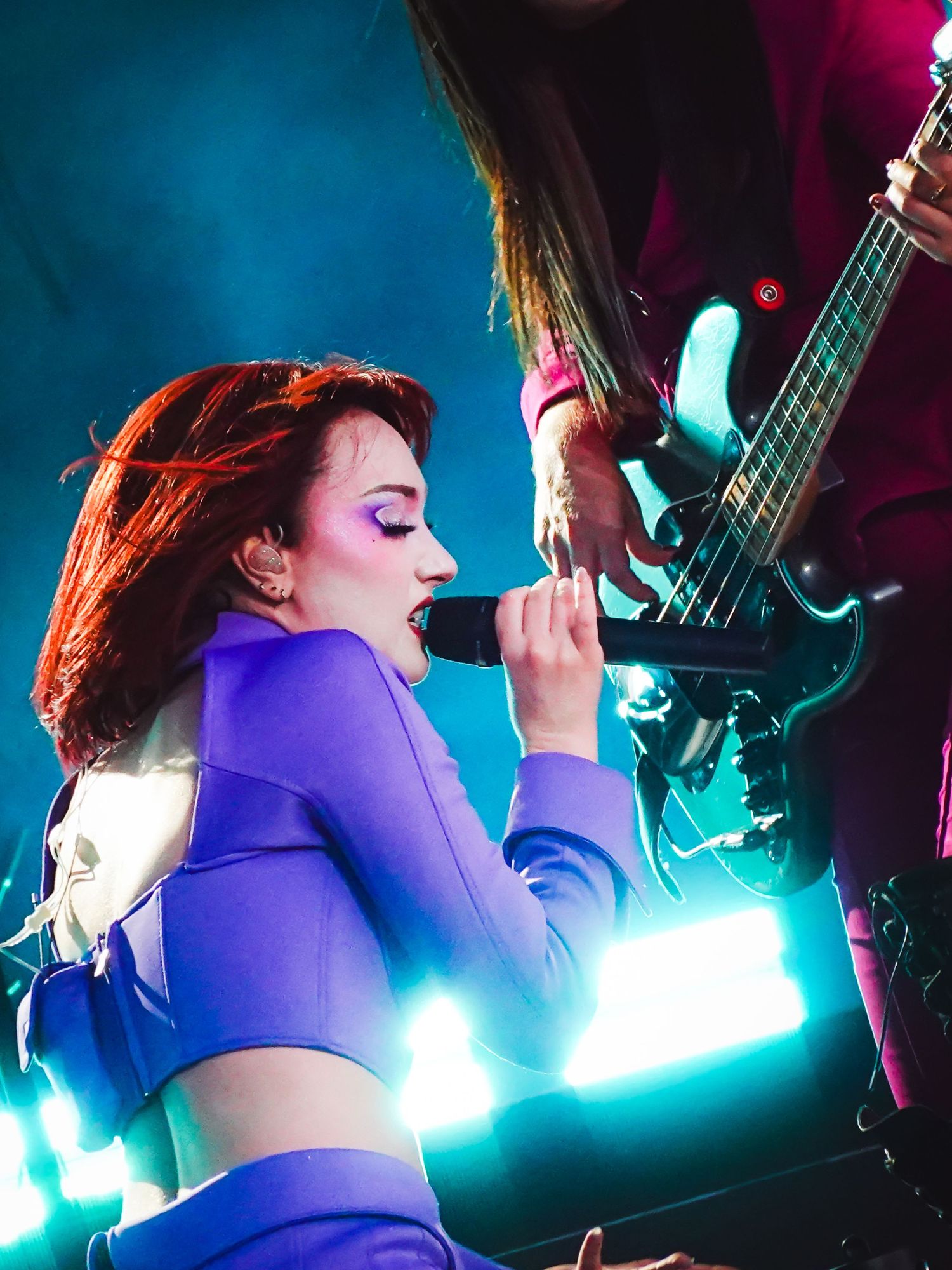 Red-haired female singer in purple crop top performs on stage with electric guitarist under blue stage lights.