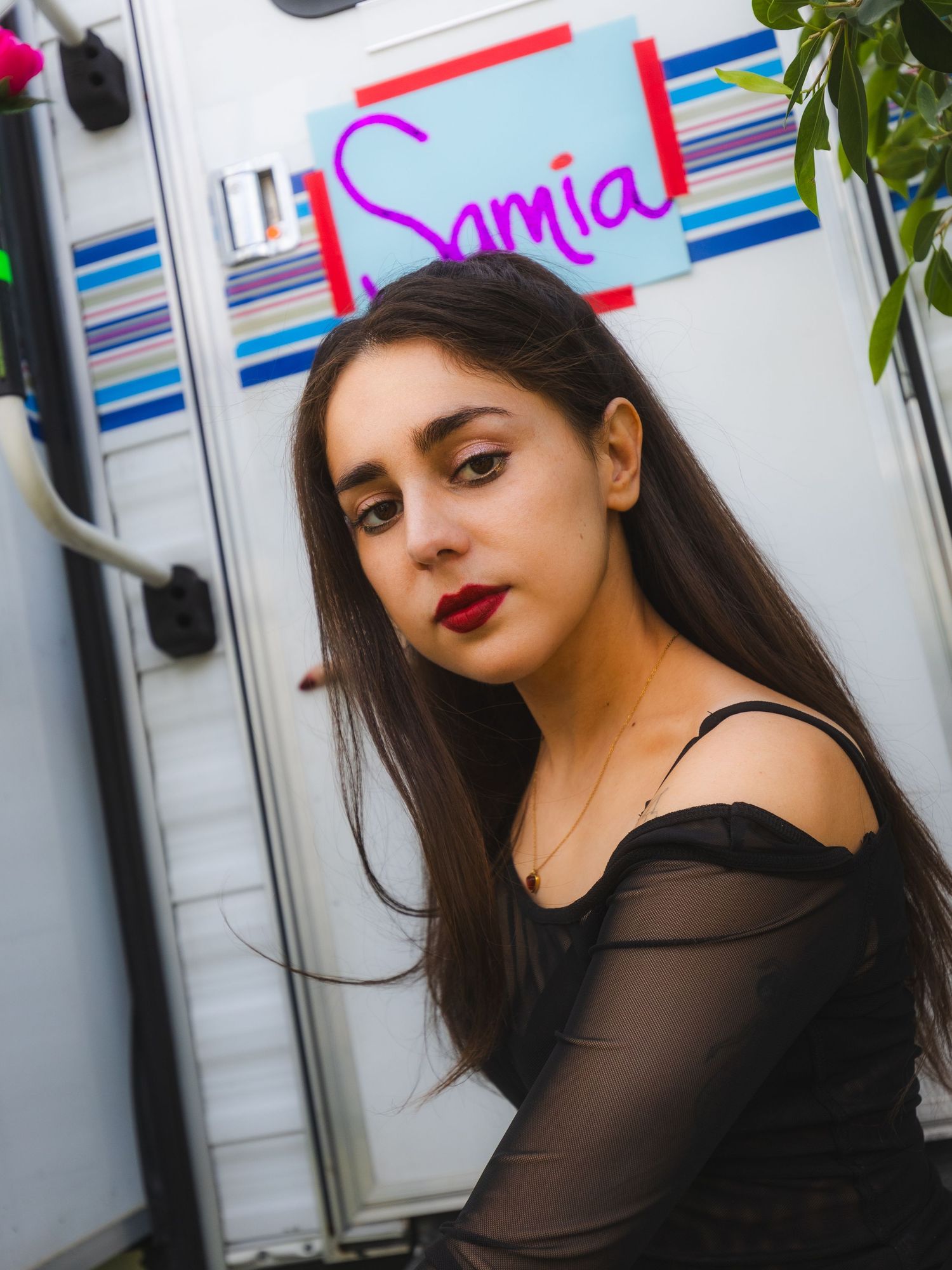 Young woman with long dark hair and red lips in black top poses near a trailer with a pink Samia sign.
