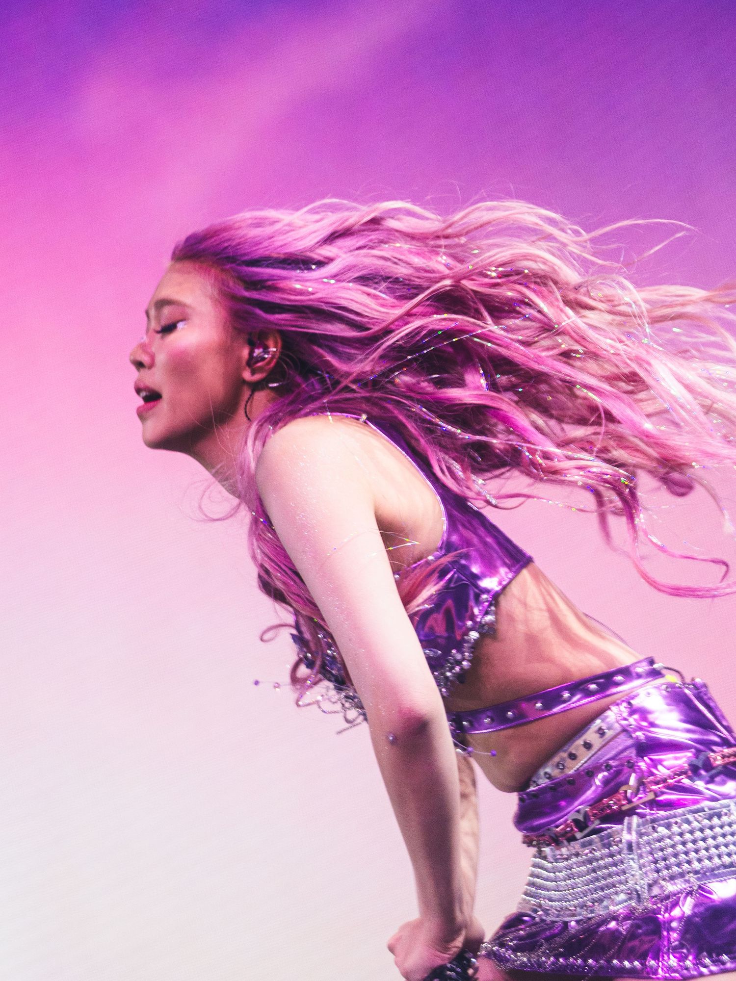 A performer with flowing pink hair dances energetically on stage in a shiny purple metallic crop top outfit.
