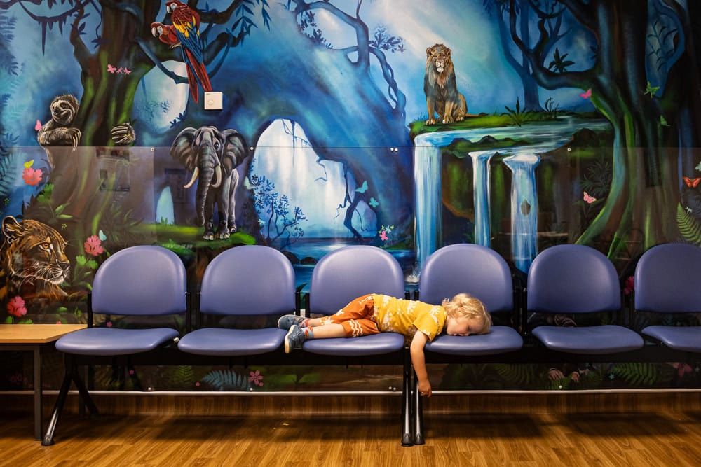 A boy pretends to fall asleep in front of a mural in a hospital A&E waiting room.