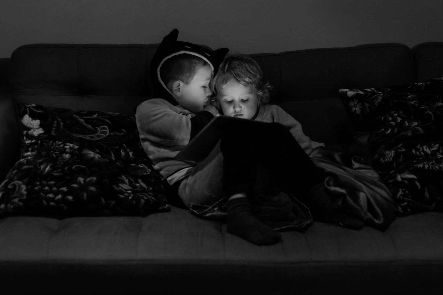 Two children sit on the sofa.  One is whispering in the other's ear.  They are lit only by the soft glow of an ipad.