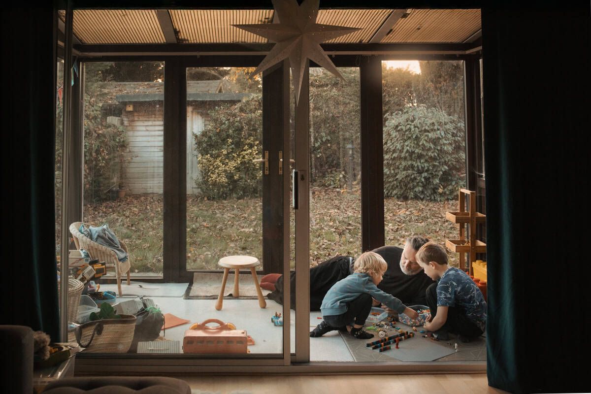 A father lies on the floor of a conservatory playroom, playing Lego with his two sons.