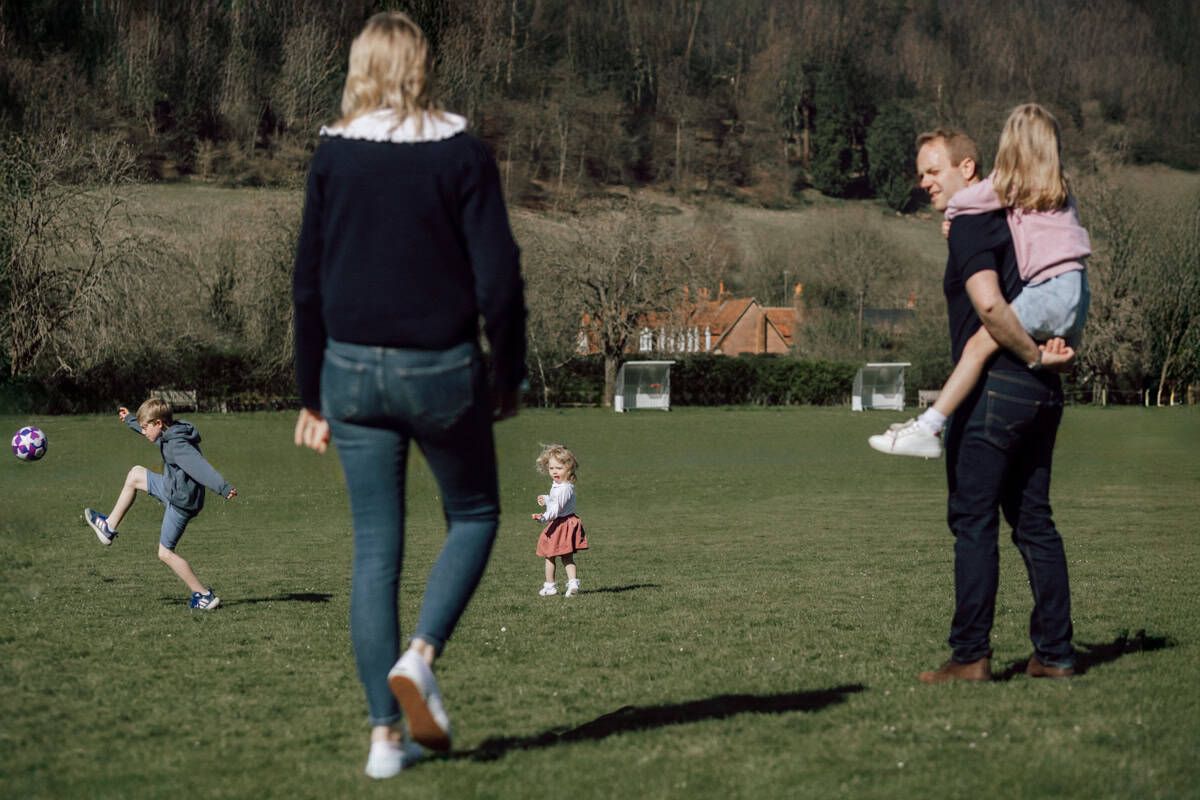 A non-traditional family portrait in which the focus rests on a boy kicking a football into the air.  HIs family are in layers around him.