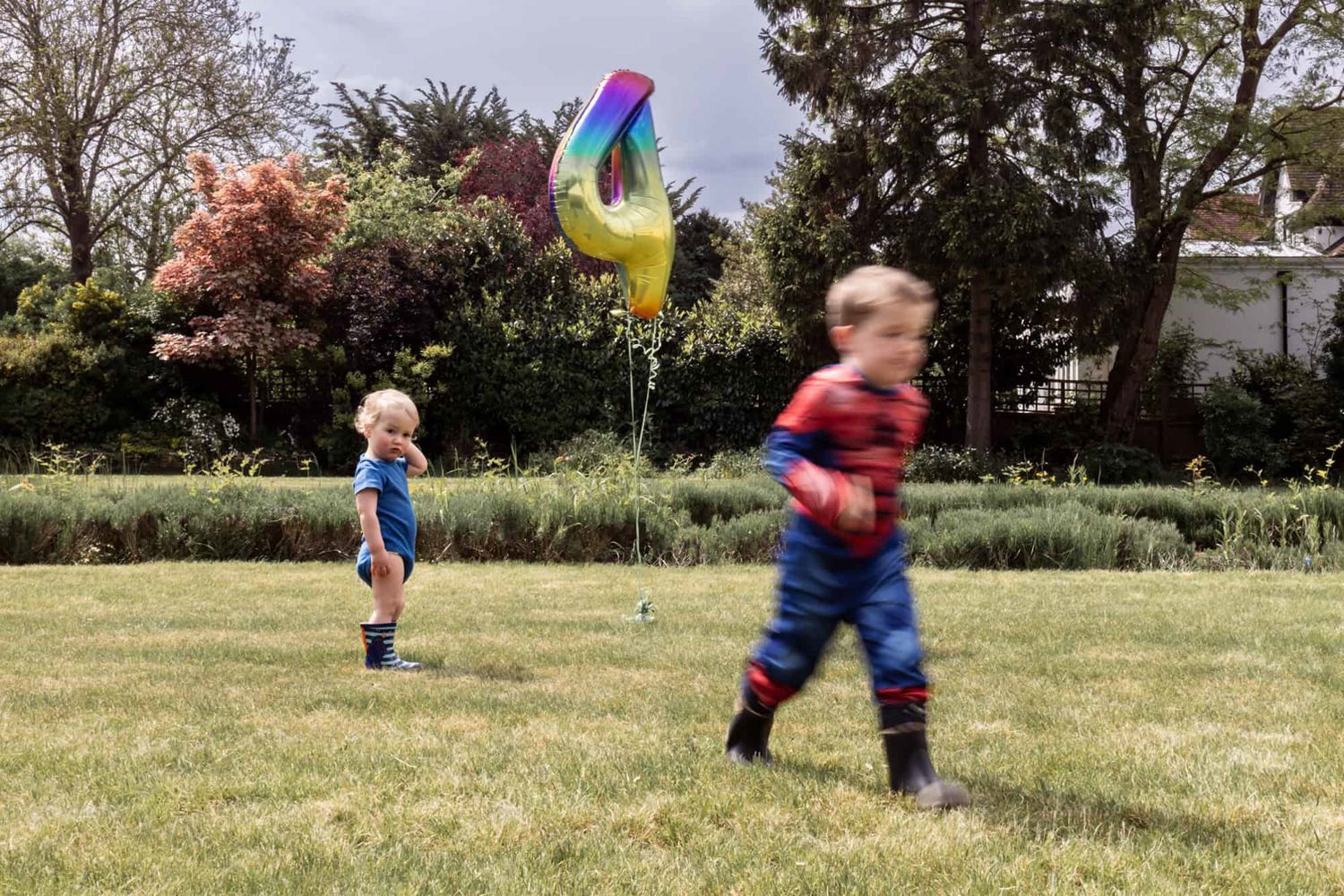 Two brothers at a birthday party.  The oldest wears a spiderman outfit and is a blur of motion, the youngest is still and confused.