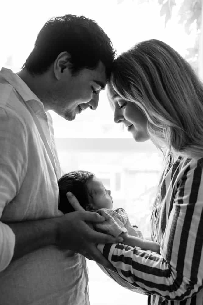 A new mother and father touch foreheads as Mum cradles their newborn between them in front of a window in the nursery.