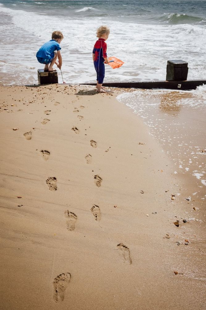 Two boys play with nets on a beach, a trail of footprints lead towards them.