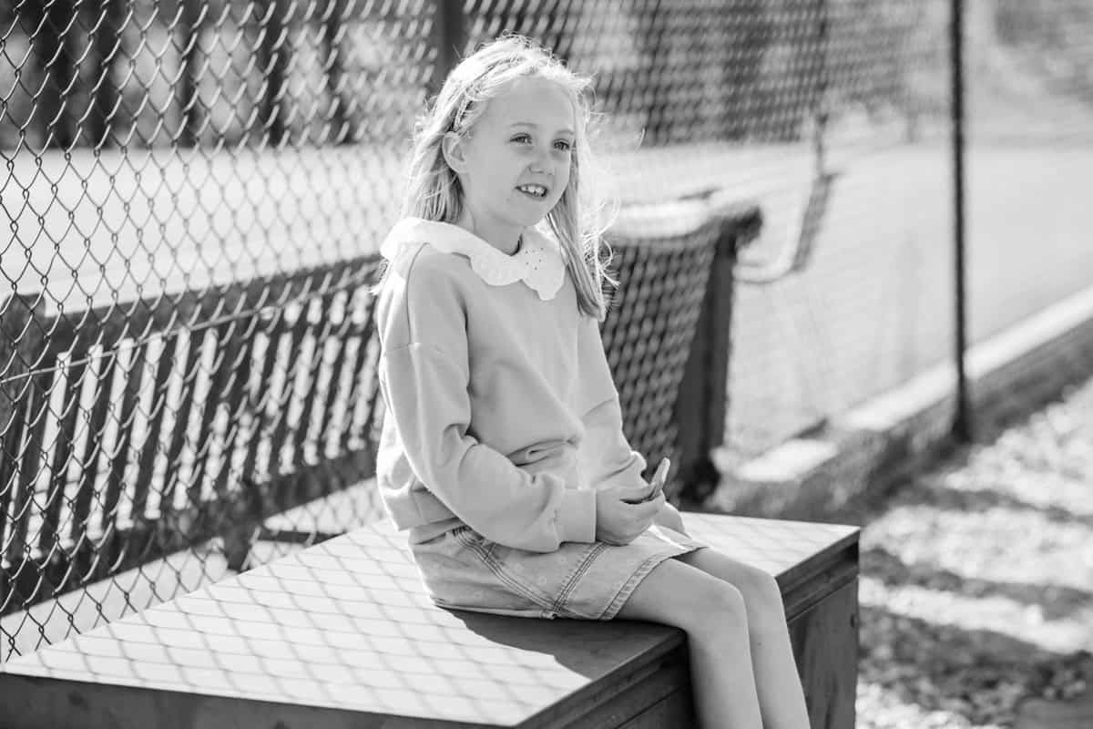 A young girl sits quietly, perched on a wooden bench in front of a fence.