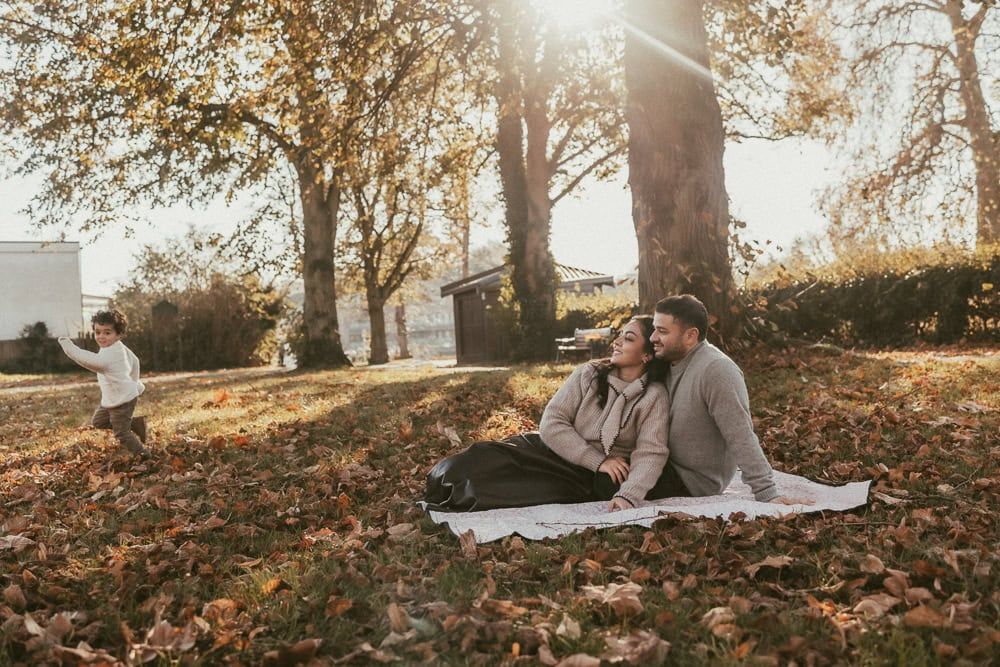 A father and mother sit together on a picnic blanket in Higginson Park, while their son runs around them.