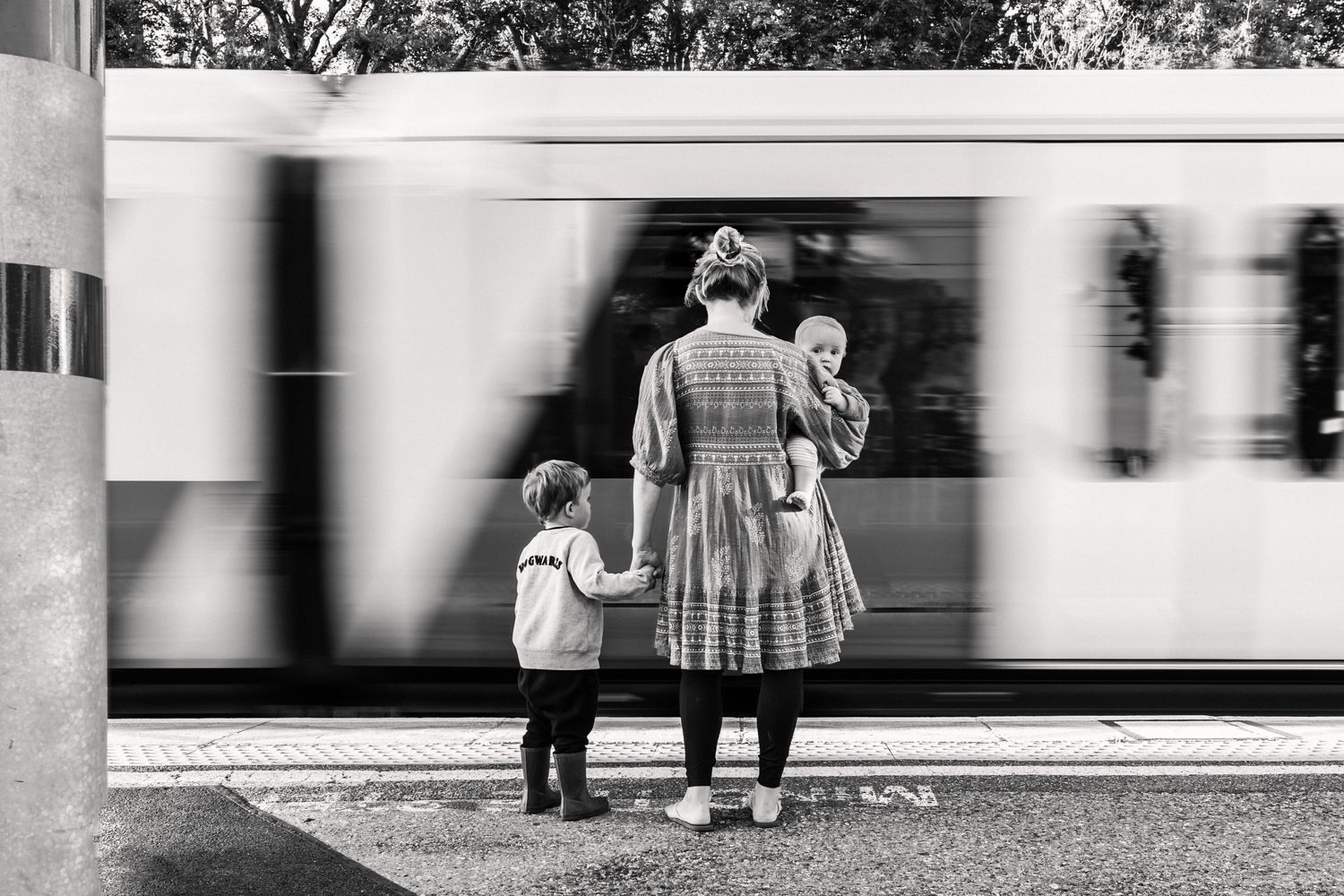 A black and white image of a mother carrying a baby and holding hands with a young boy as a moving train whizzes past.