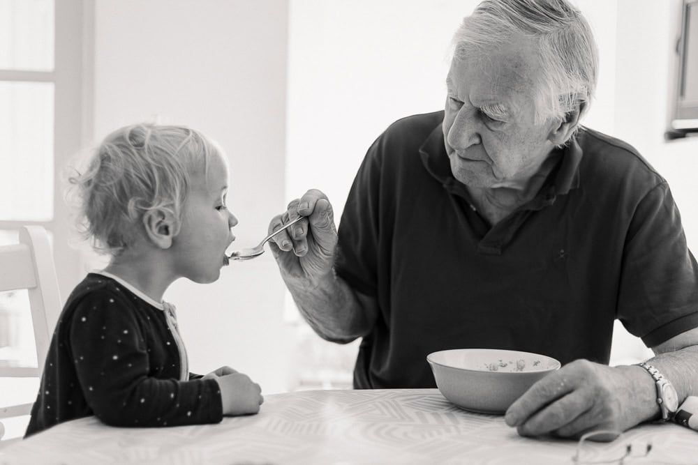 A grandfather feeds his grandson from his breakfast bowl.