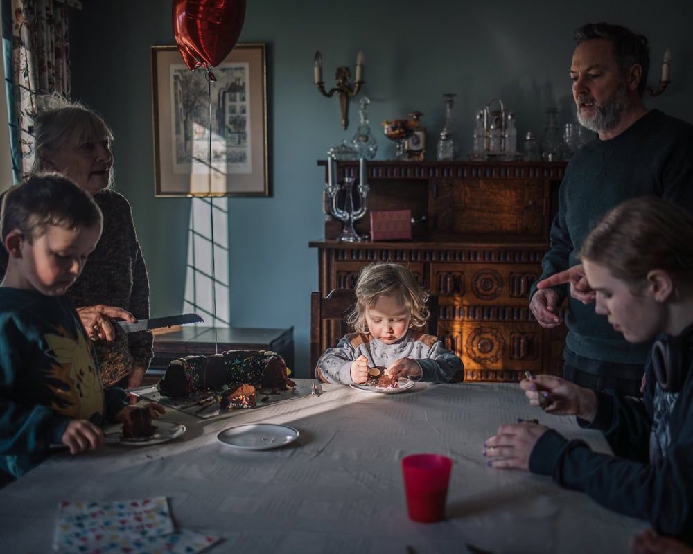 A boy sits at the head of a dining table tucking under his birthday cake.  He is surrounded by close family.